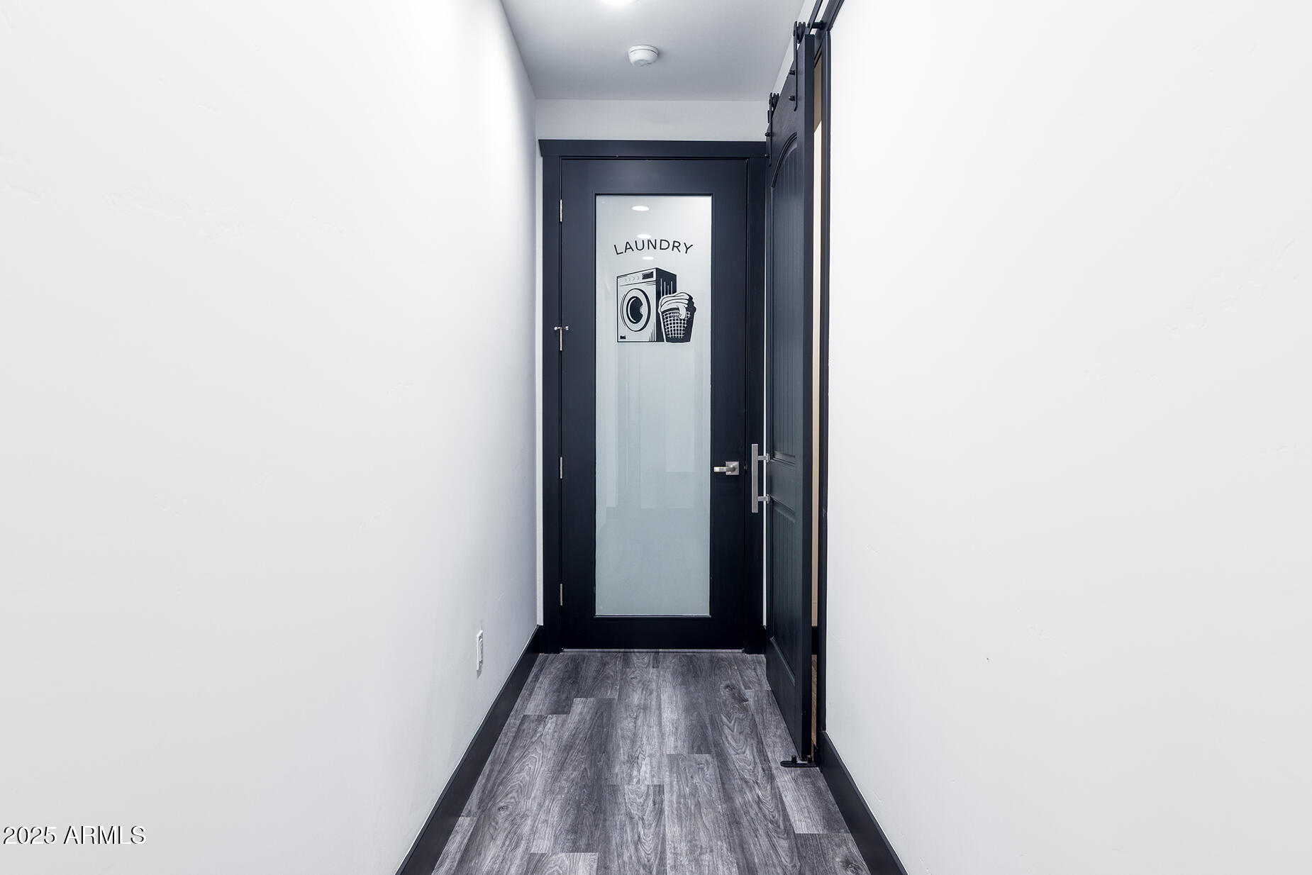 Hallway leading to a laundry room, with a black-framed frosted-glass door and gray plank flooring.