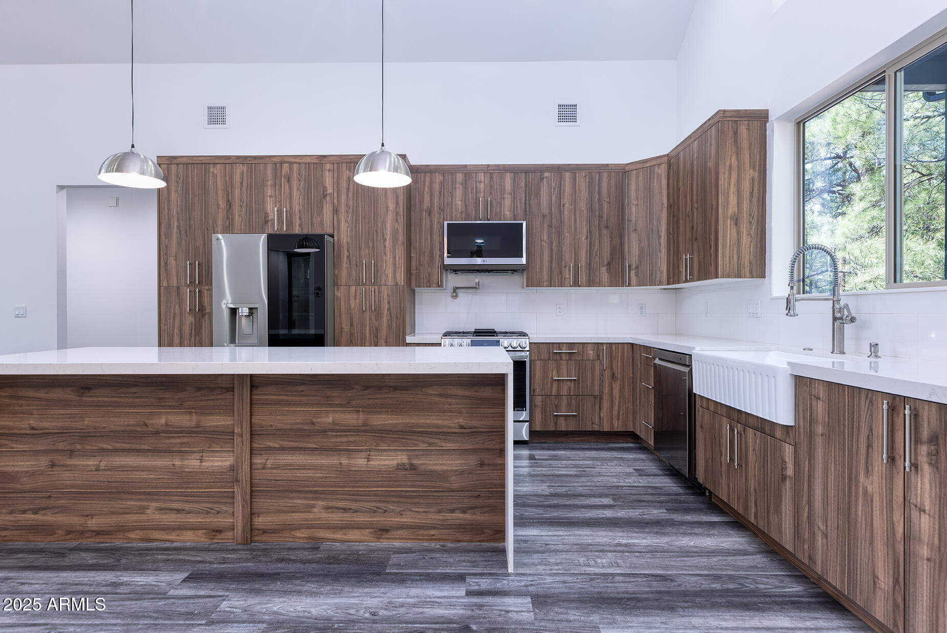 Modern kitchen with walnut cabinetry, a large white-countertop island, stainless appliances, and a farmhouse sink by wide windows.