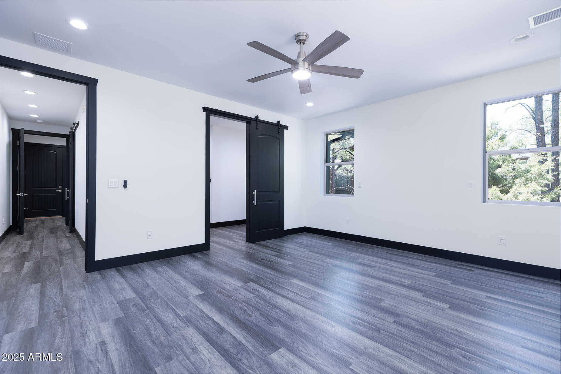 Vacant room with gray plank flooring, black barn door, ceiling fan, and windows overlooking trees.