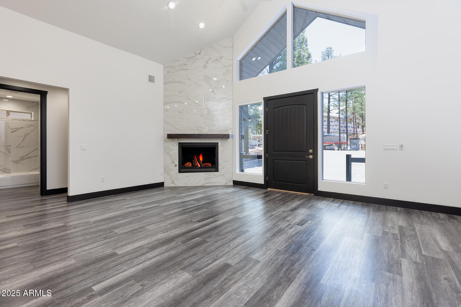 Vacant living room with a tall marble-look fireplace, gray flooring, and high windows overlooking pine trees.