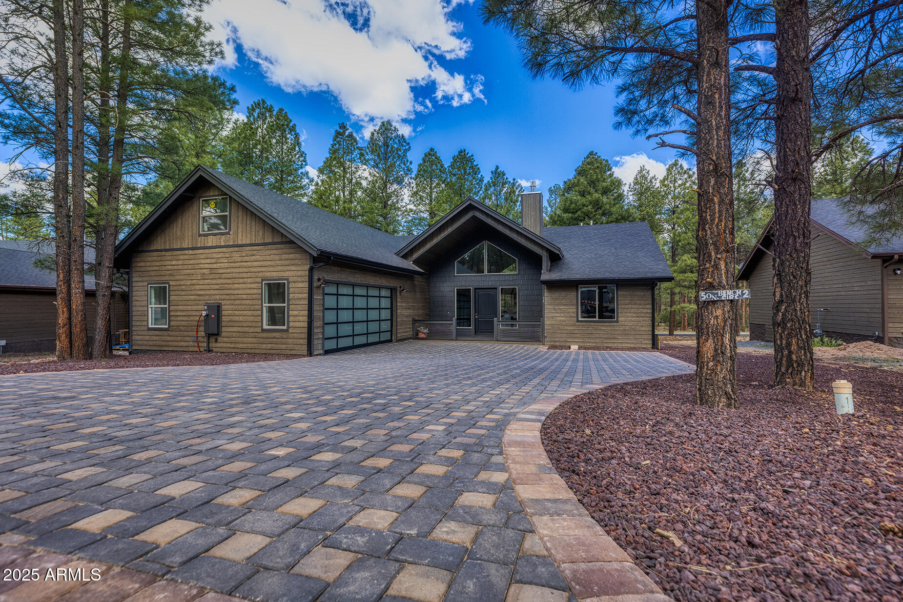 Modern wood-sided home with a glass-panel garage door, wide paver driveway, and tall pine trees.