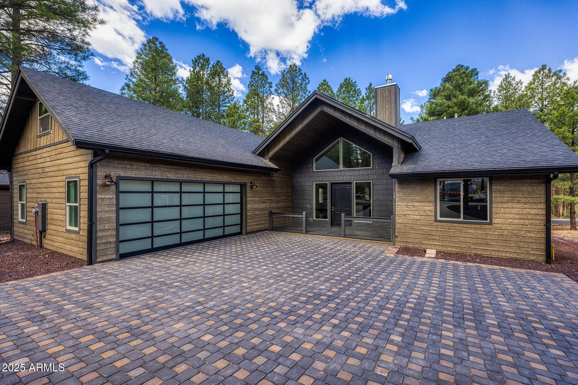 Modern wood-sided home with a glass-panel garage door and broad paved driveway among pine trees.