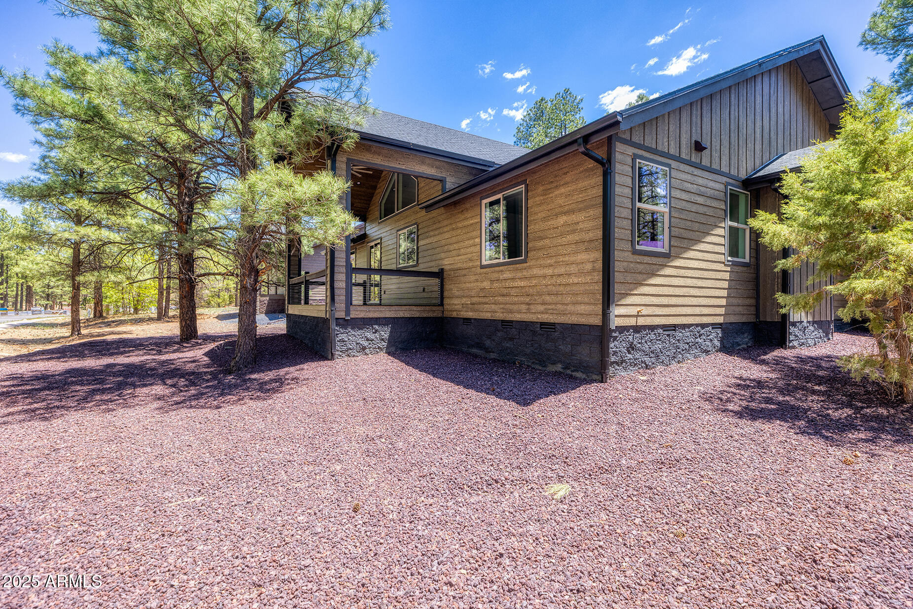 Wood-sided home with a covered porch among pine trees on a gravel-covered lot.