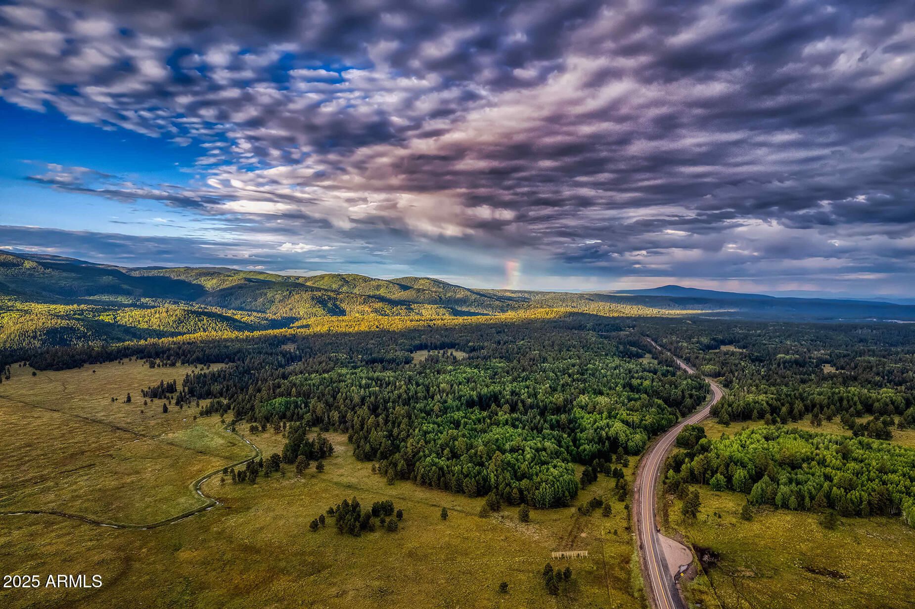 Aerial view of forested hills and meadow, with a curving road and a faint rainbow beneath dramatic clouds.