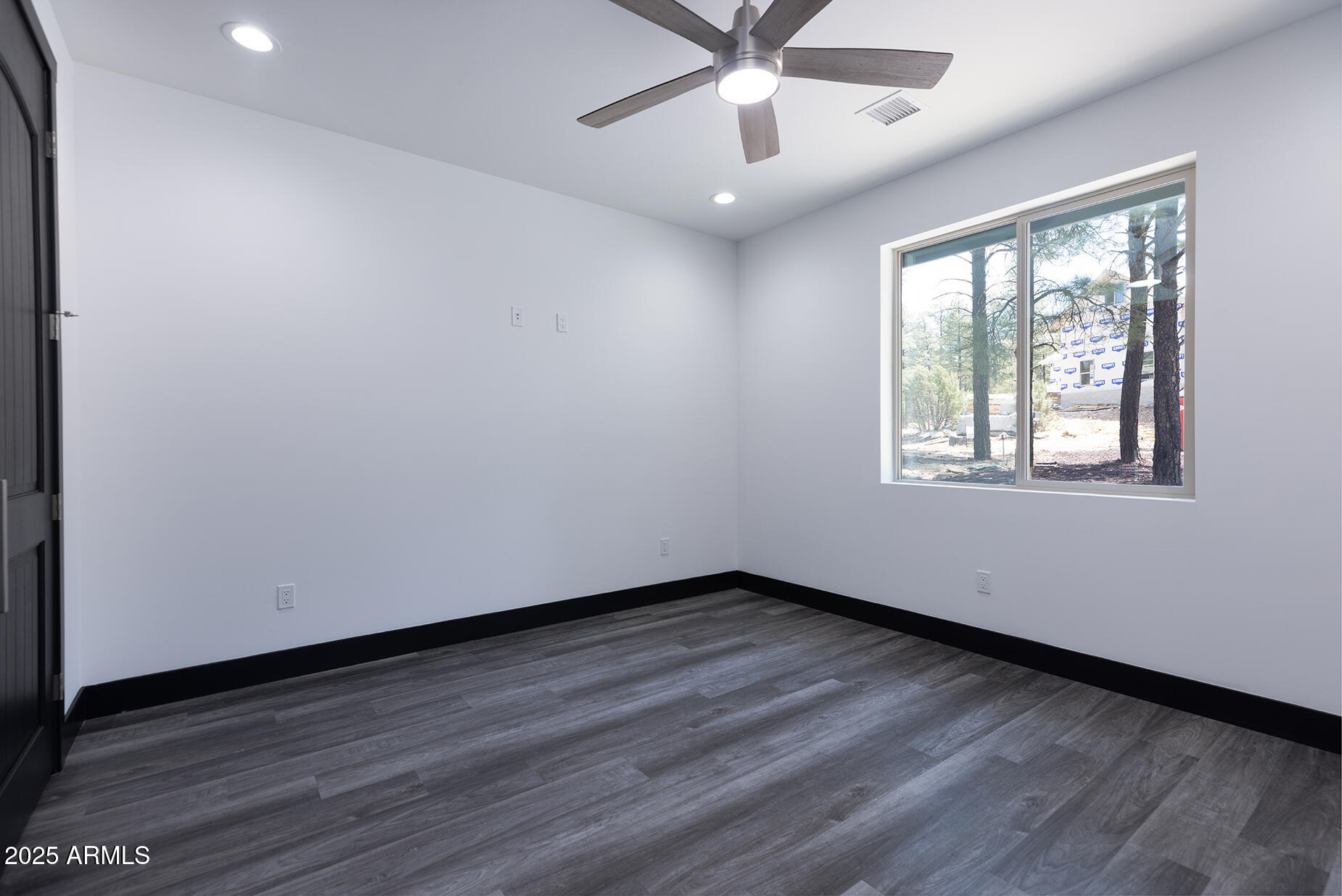 Empty bedroom with gray plank flooring, a ceiling fan, and a large window overlooking trees.