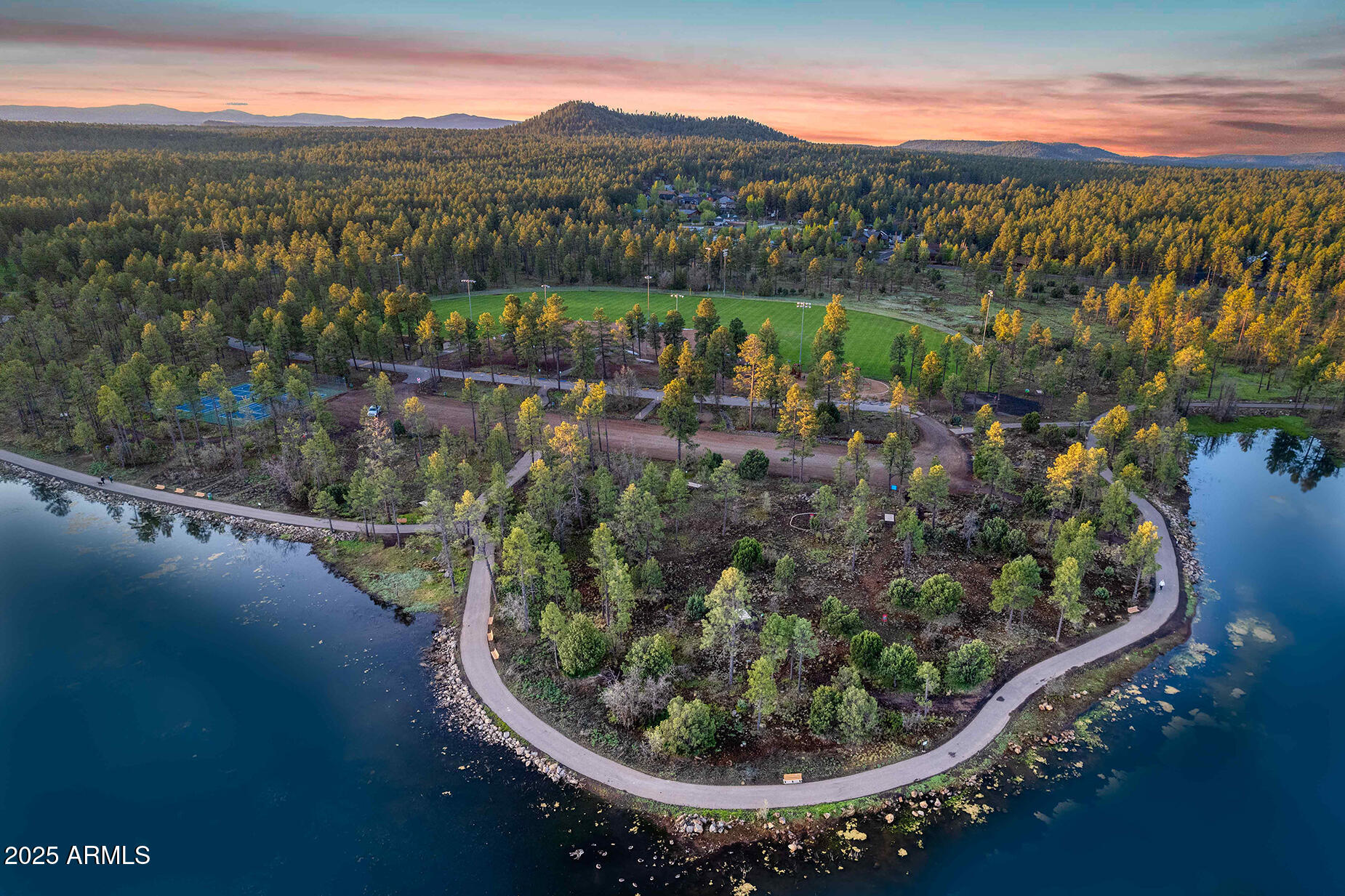 Aerial lakeside park with winding path, sports fields, and forested hills at sunset.
