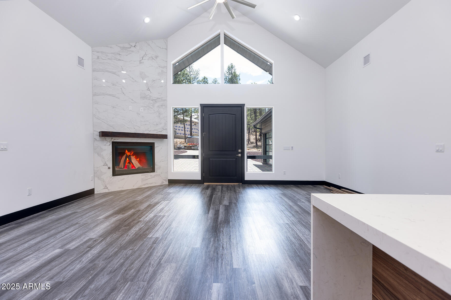 Vaulted great room with a marble-look fireplace, black front door, and tall windows facing pine trees.