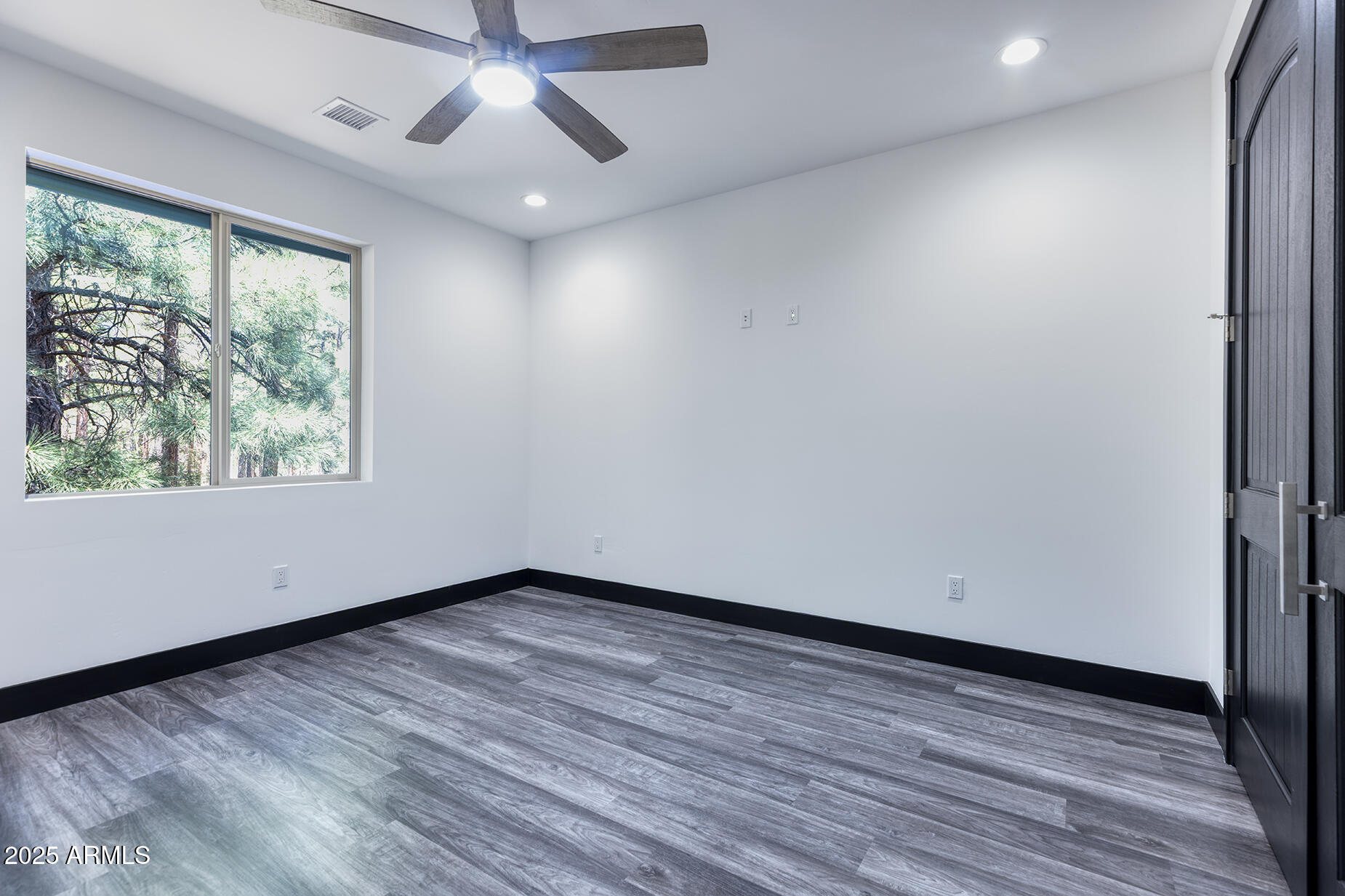Empty room with gray wood-look flooring, a ceiling fan, and a window overlooking pine trees.