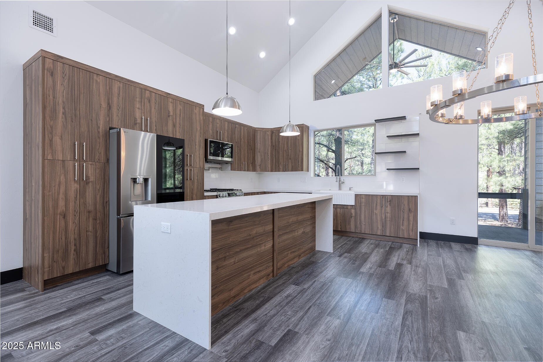Modern kitchen with walnut cabinets, a large white island, stainless steel appliances, and windows overlooking pine trees.
