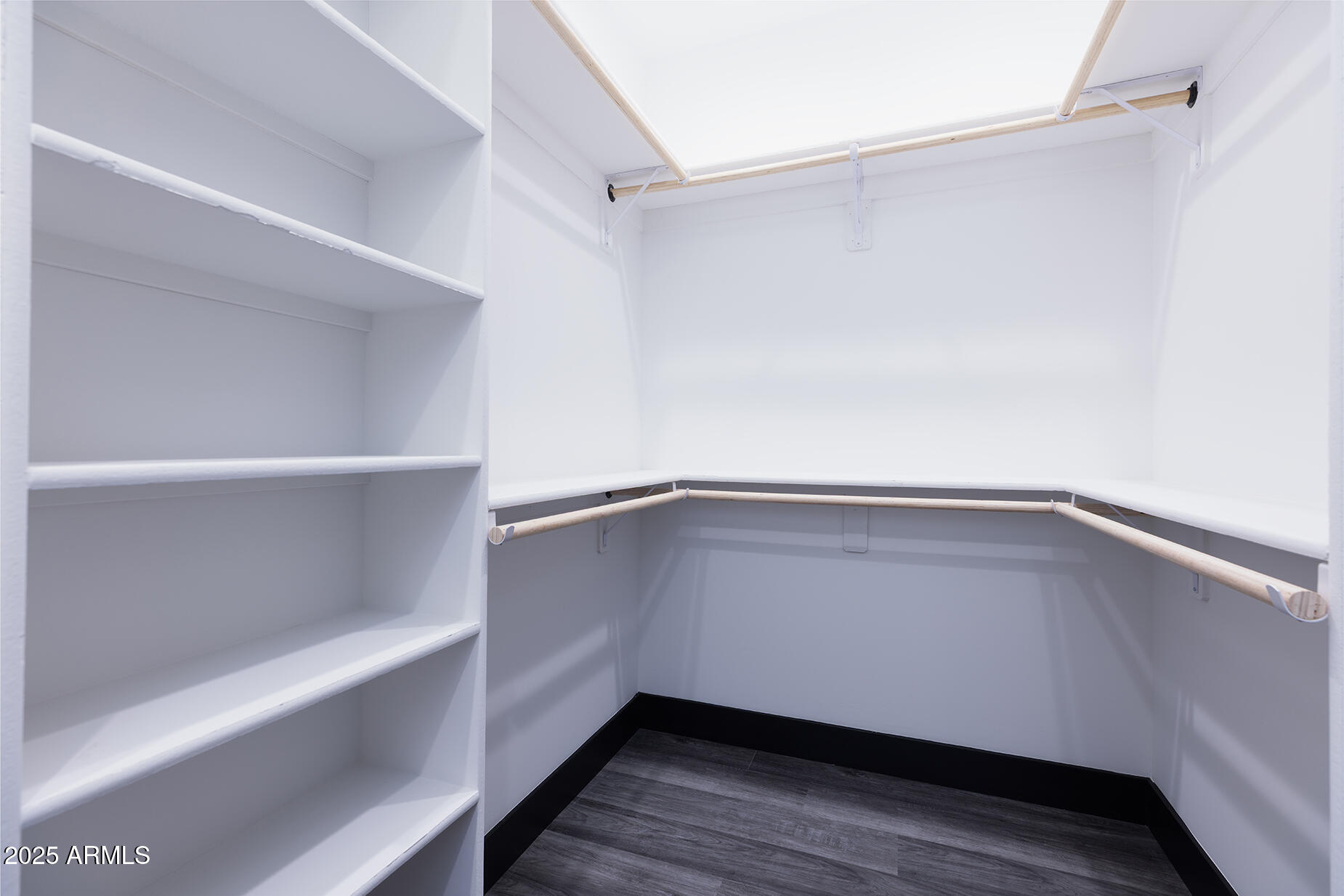 Walk-in closet with white built-in shelves, wraparound clothes rods, and dark flooring.