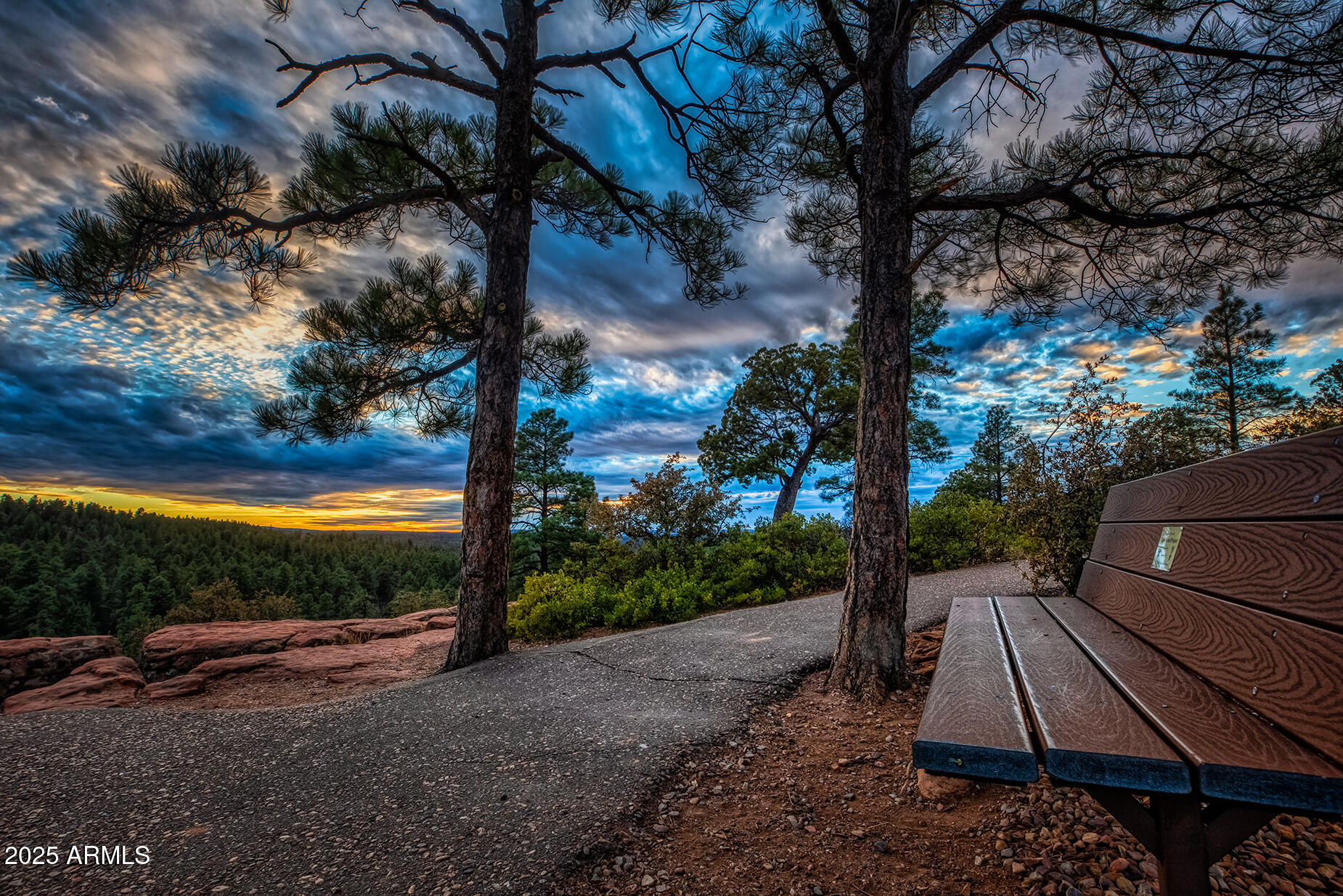 Forest overlook with a wooden bench beside a paved path beneath a dramatic sunset sky.