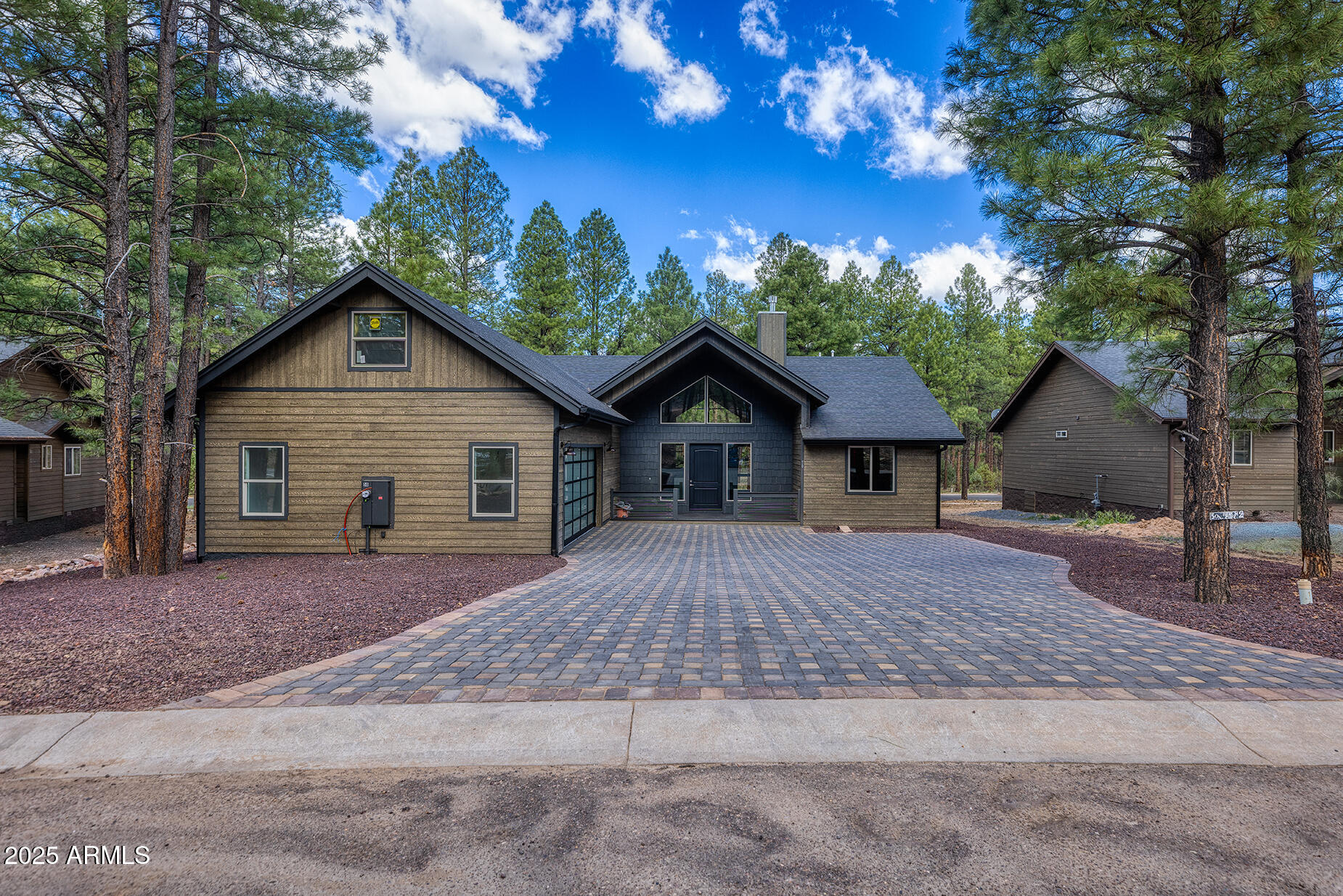 Wood-sided home with a paver driveway, glass-paneled garage door, and tall pines.