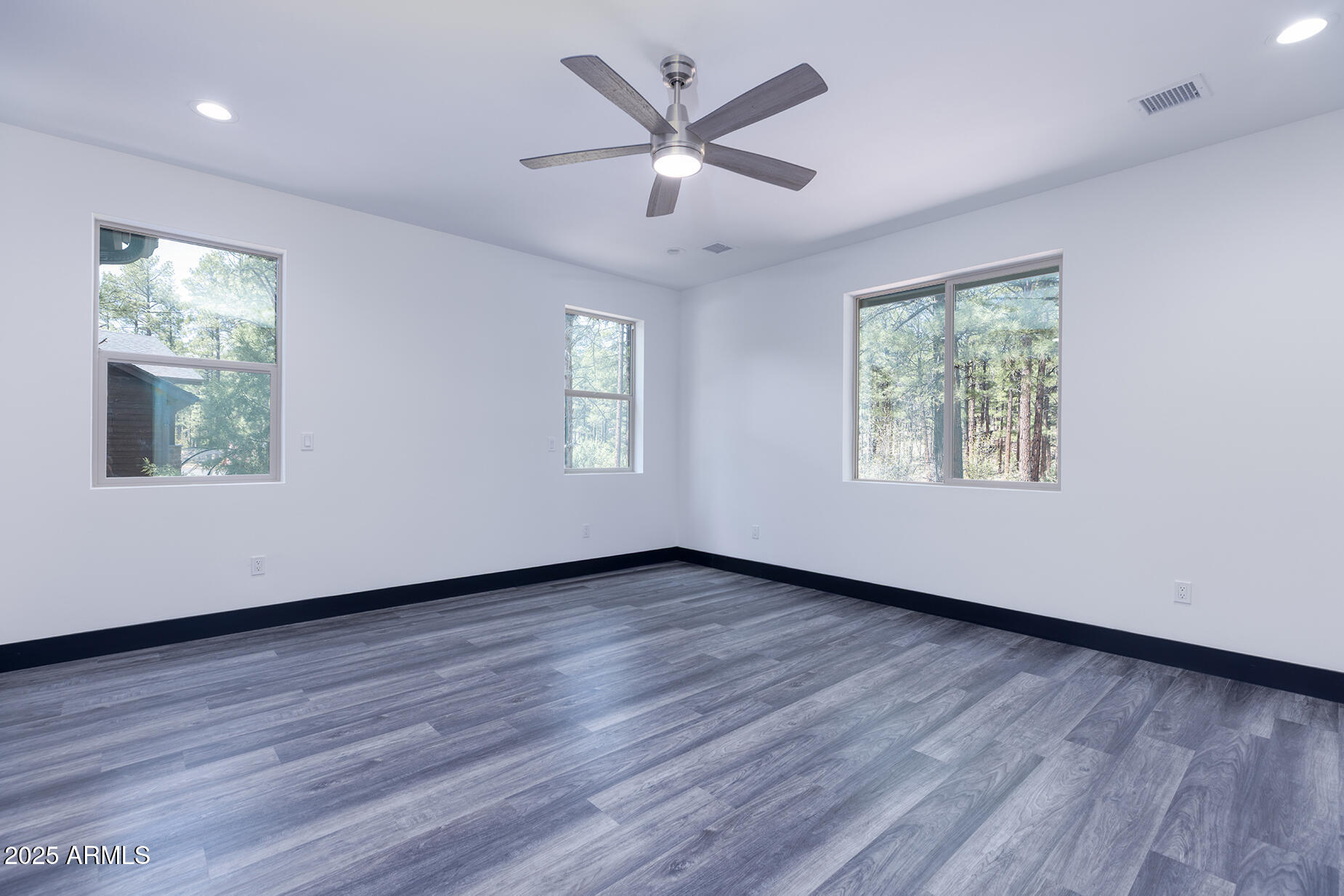 Empty room with gray wood-look flooring, a ceiling fan, and windows overlooking trees.