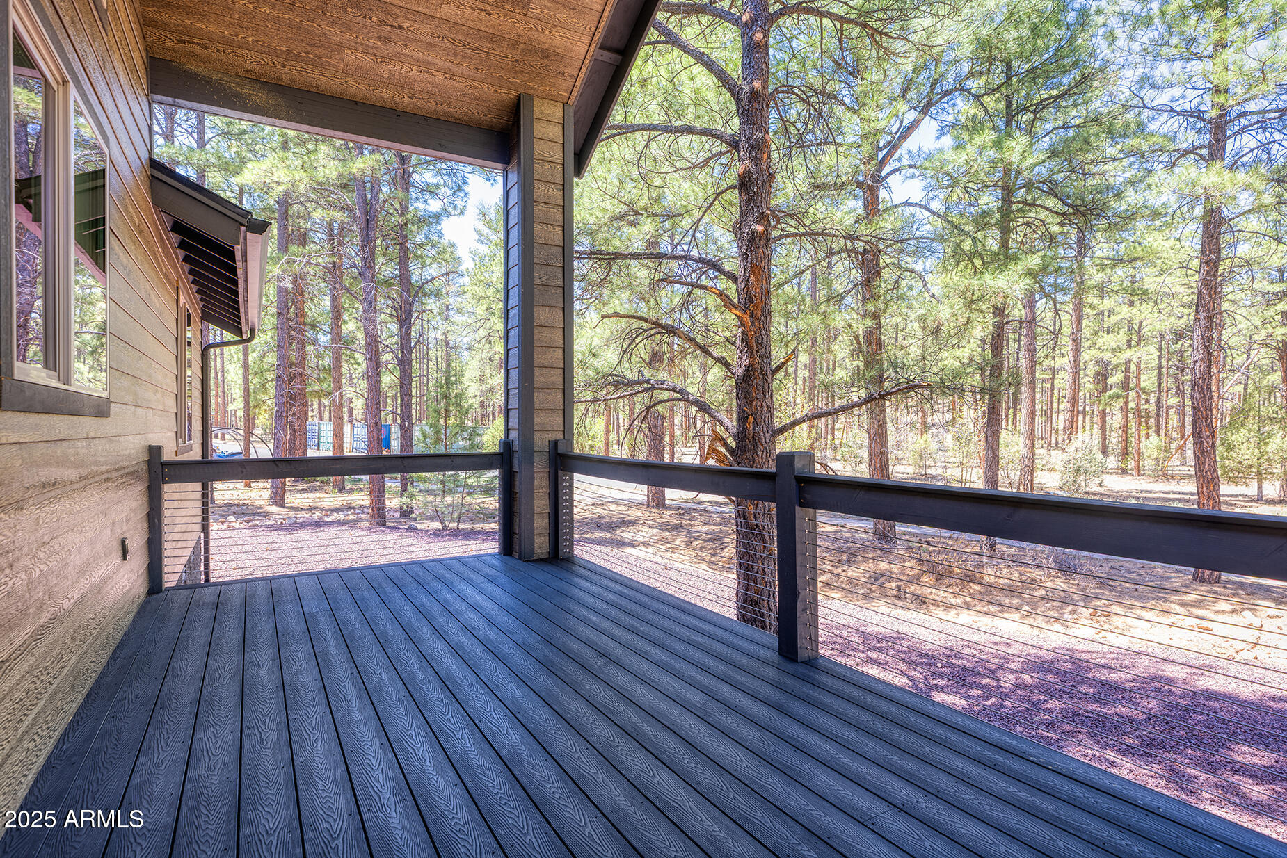 Covered wooden deck with cable railing overlooking a sunlit pine forest.