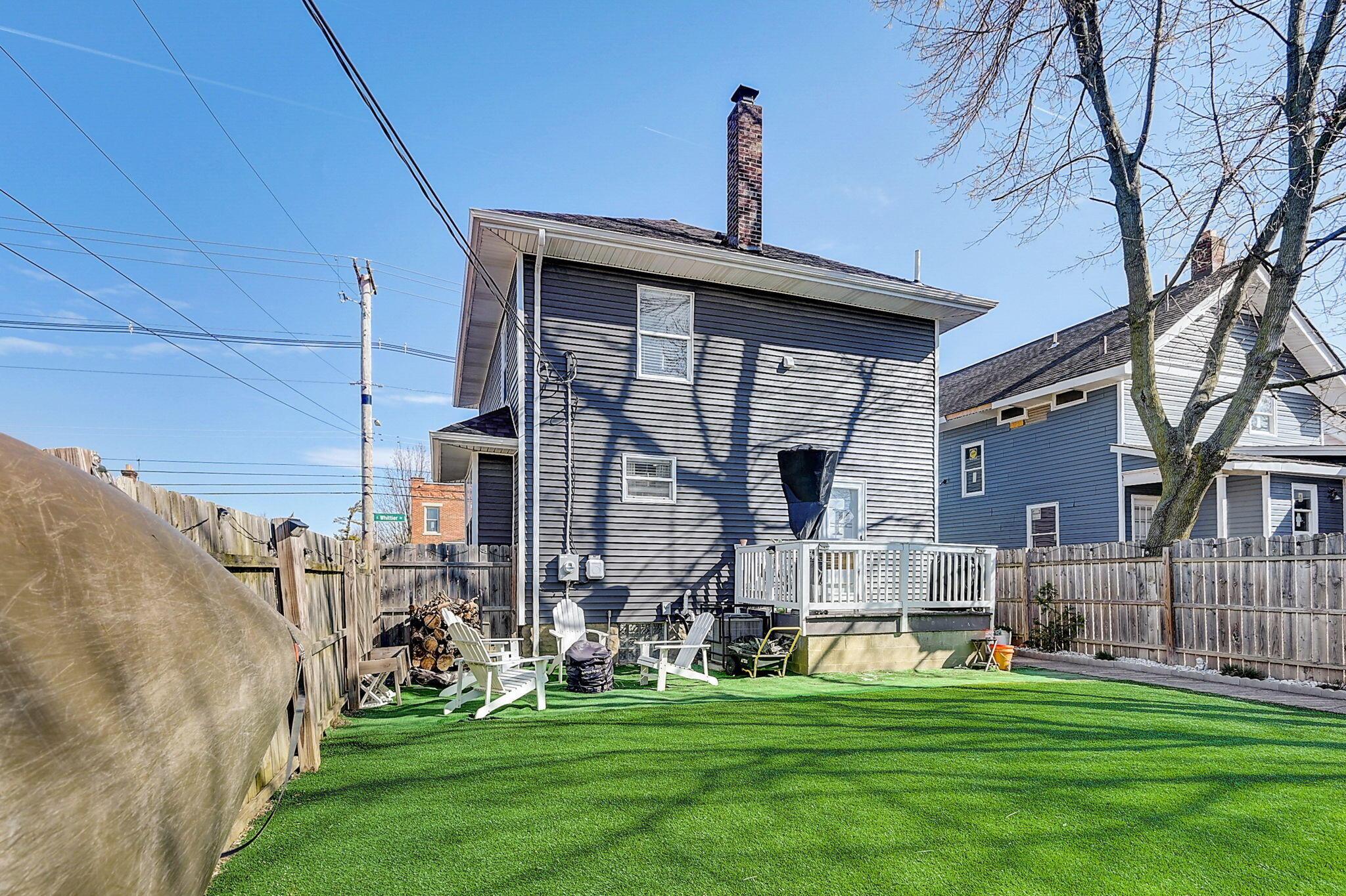 Fenced backyard with artificial turf, outdoor seating, and a raised deck behind a gray-sided house.