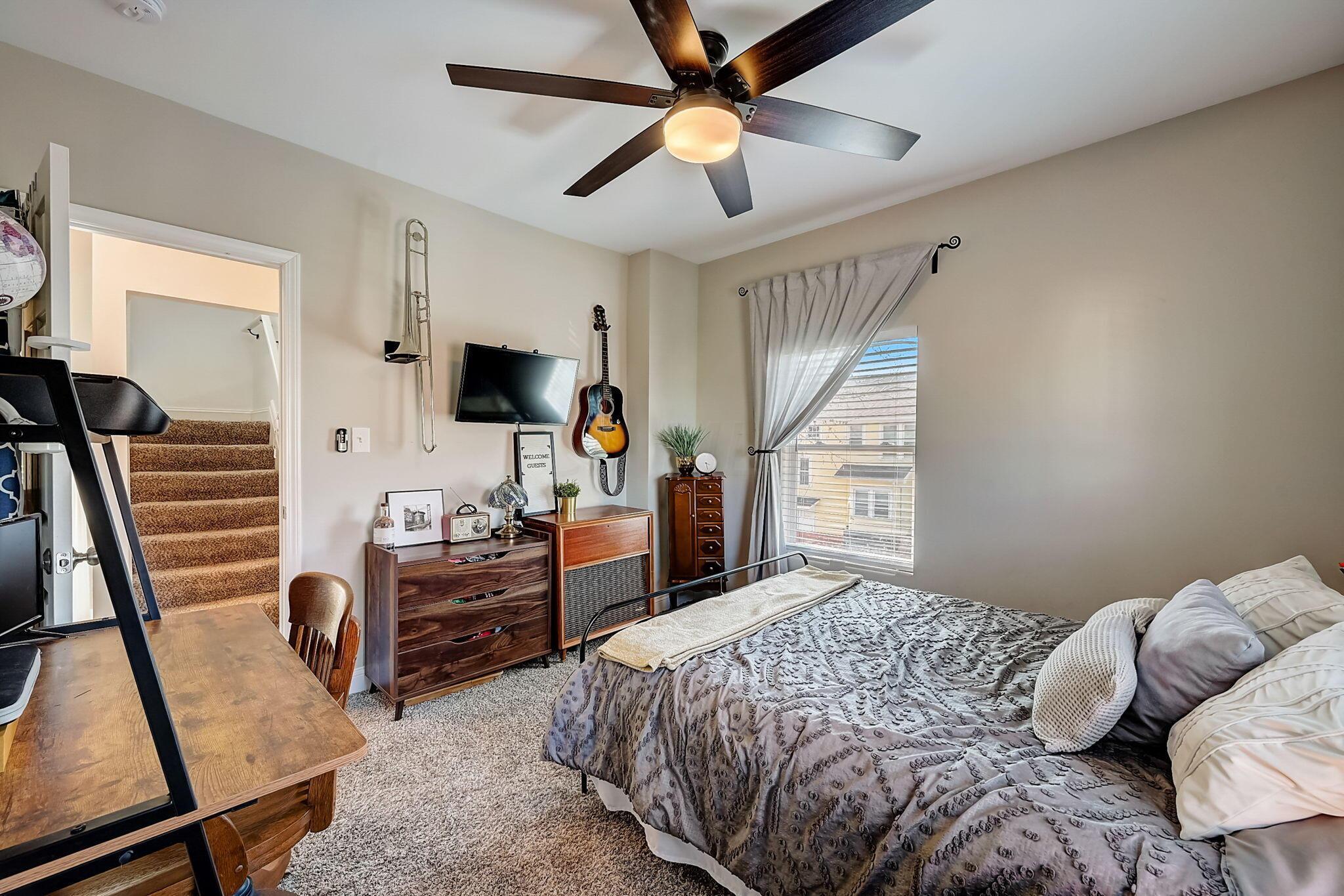 Furnished bedroom with a patterned gray bedspread, wooden dressers, a wall-mounted guitar, and a carpeted staircase.