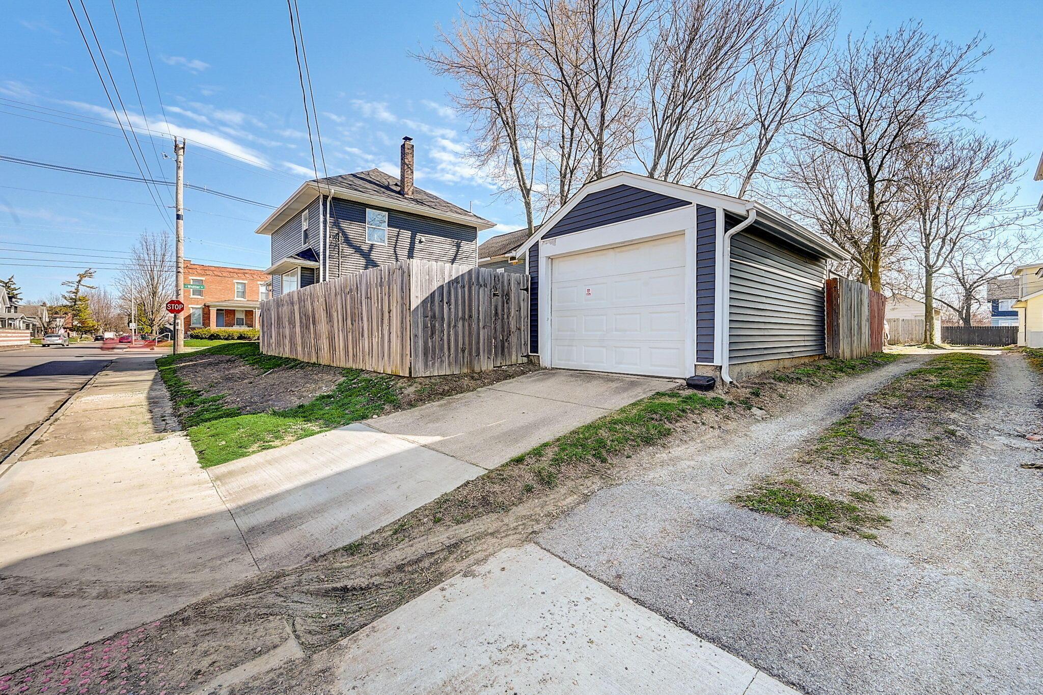 Detached blue-gray garage with a white door beside a wooden fence and gravel alley.