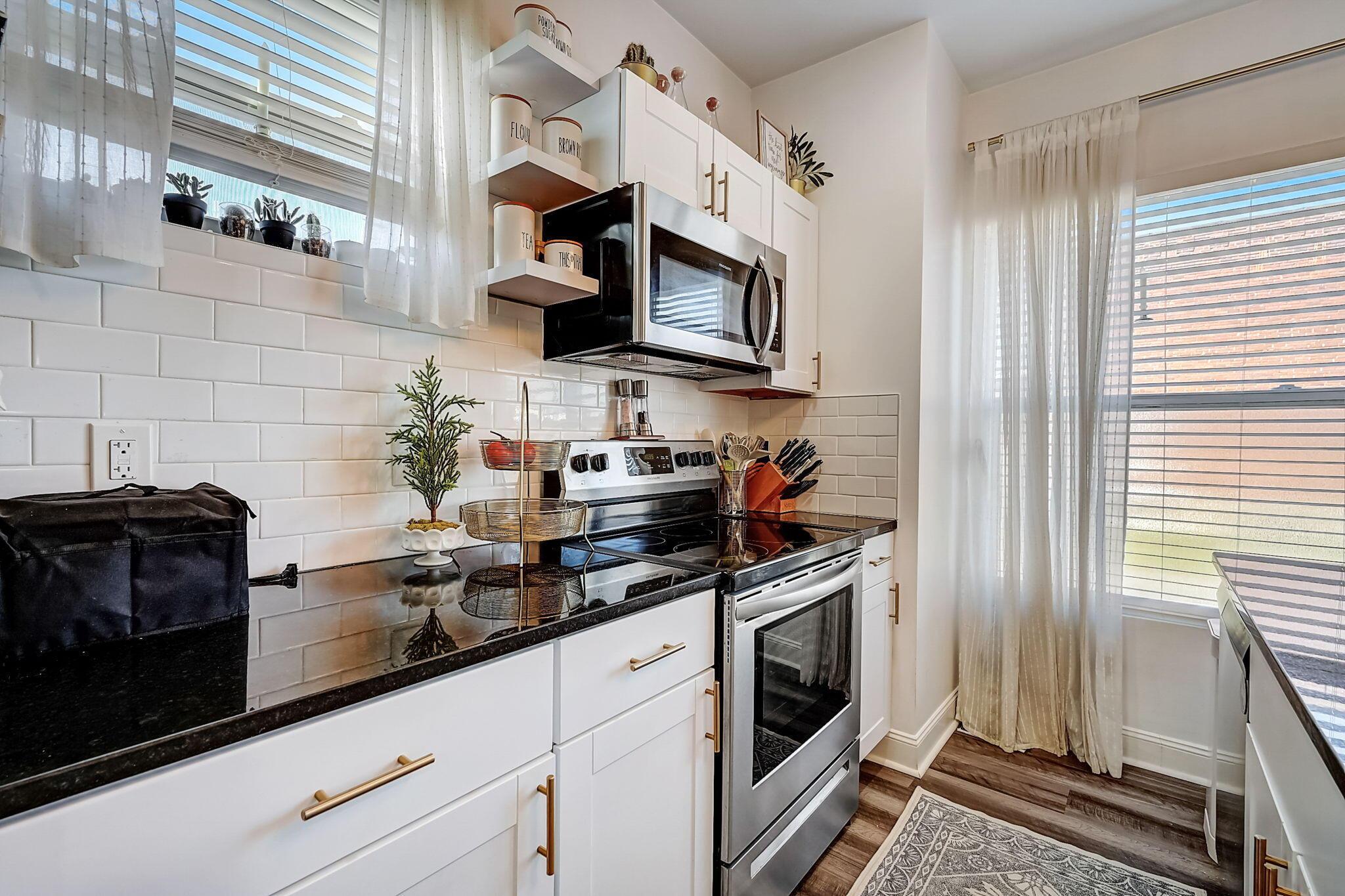 Bright kitchen with white cabinets, black counters, subway tile, and stainless steel appliances beside large windows.