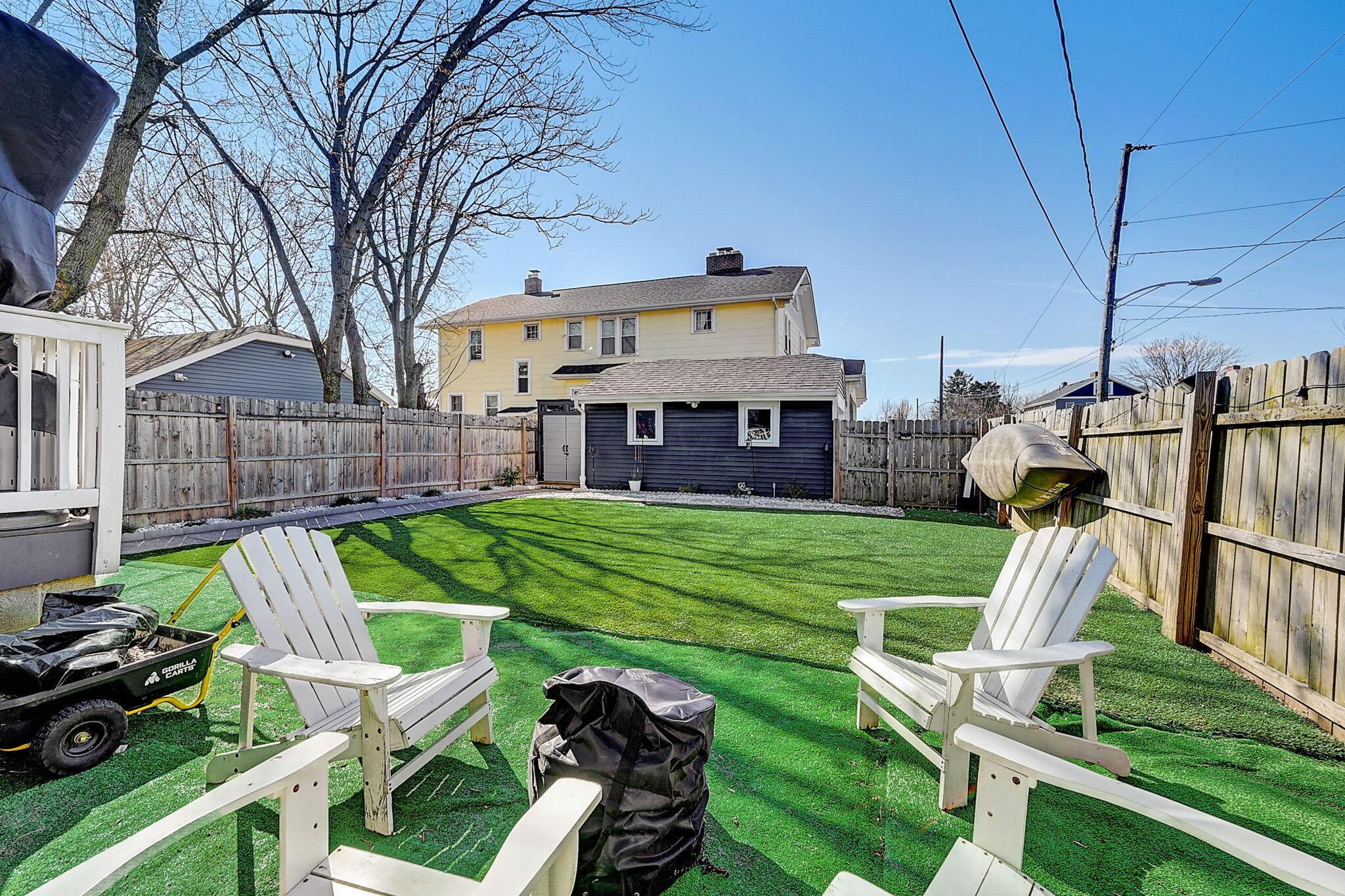 Fenced backyard with artificial turf, white Adirondack chairs, and the rear of the house.