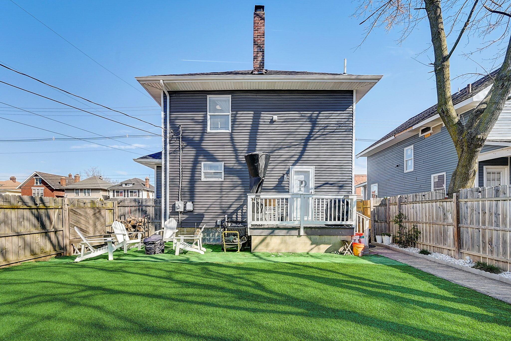 Fenced backyard with artificial turf, Adirondack chairs, and a small deck behind a dark-sided house.