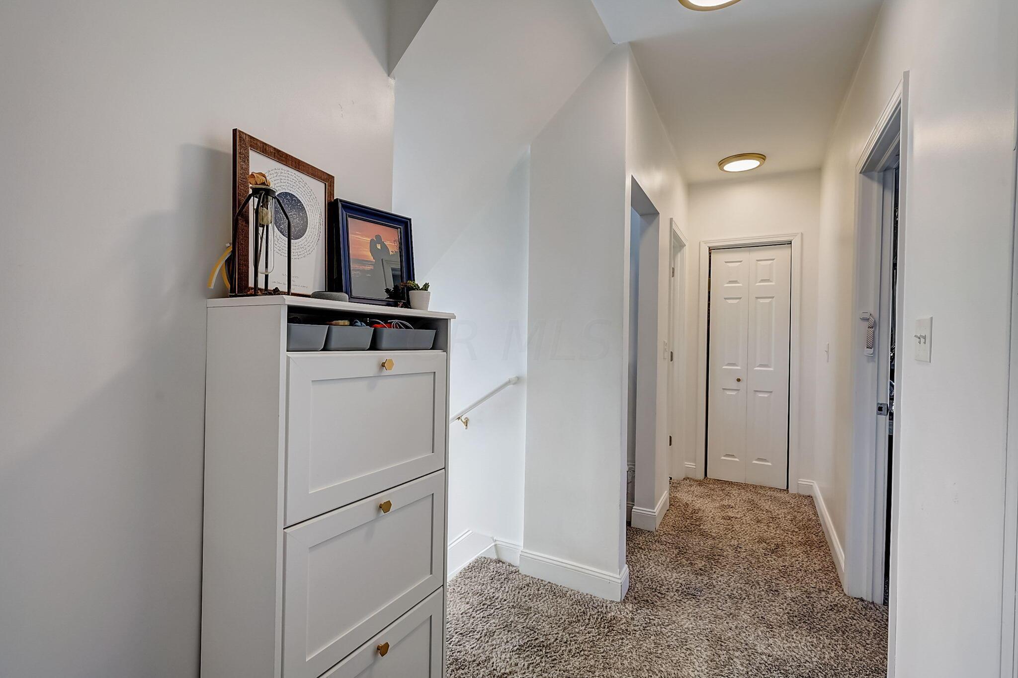 Carpeted upstairs hallway with double closet doors and a white cabinet topped with framed artwork.