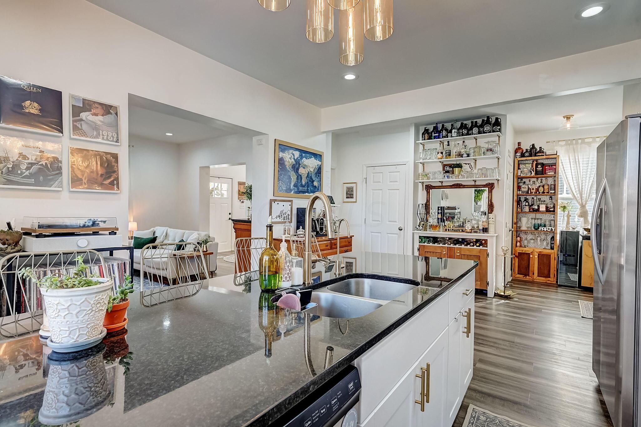 Open-plan kitchen with a dark stone island, large sink, brass-accented cabinets, and stocked bar shelves.