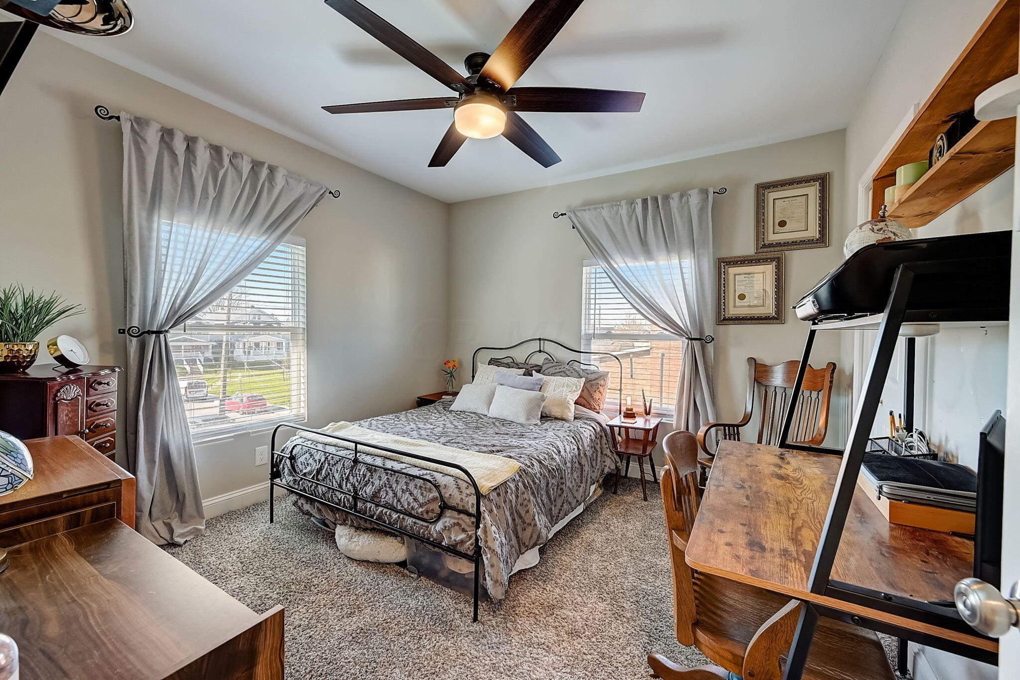 Bedroom with a metal-frame bed, patterned gray bedding, curtained windows, and a wooden desk.