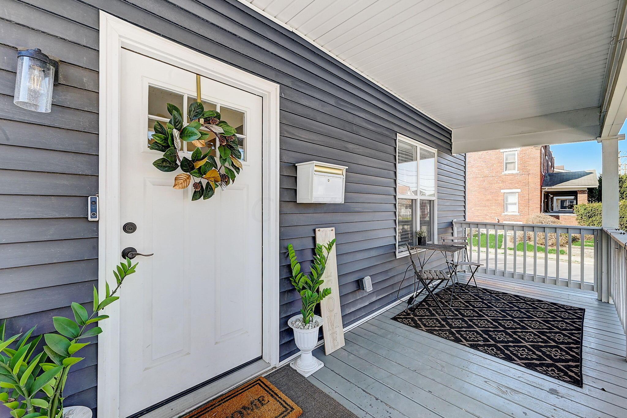 Covered front porch with a white wreath-decorated door, potted plants, and bistro seating.
