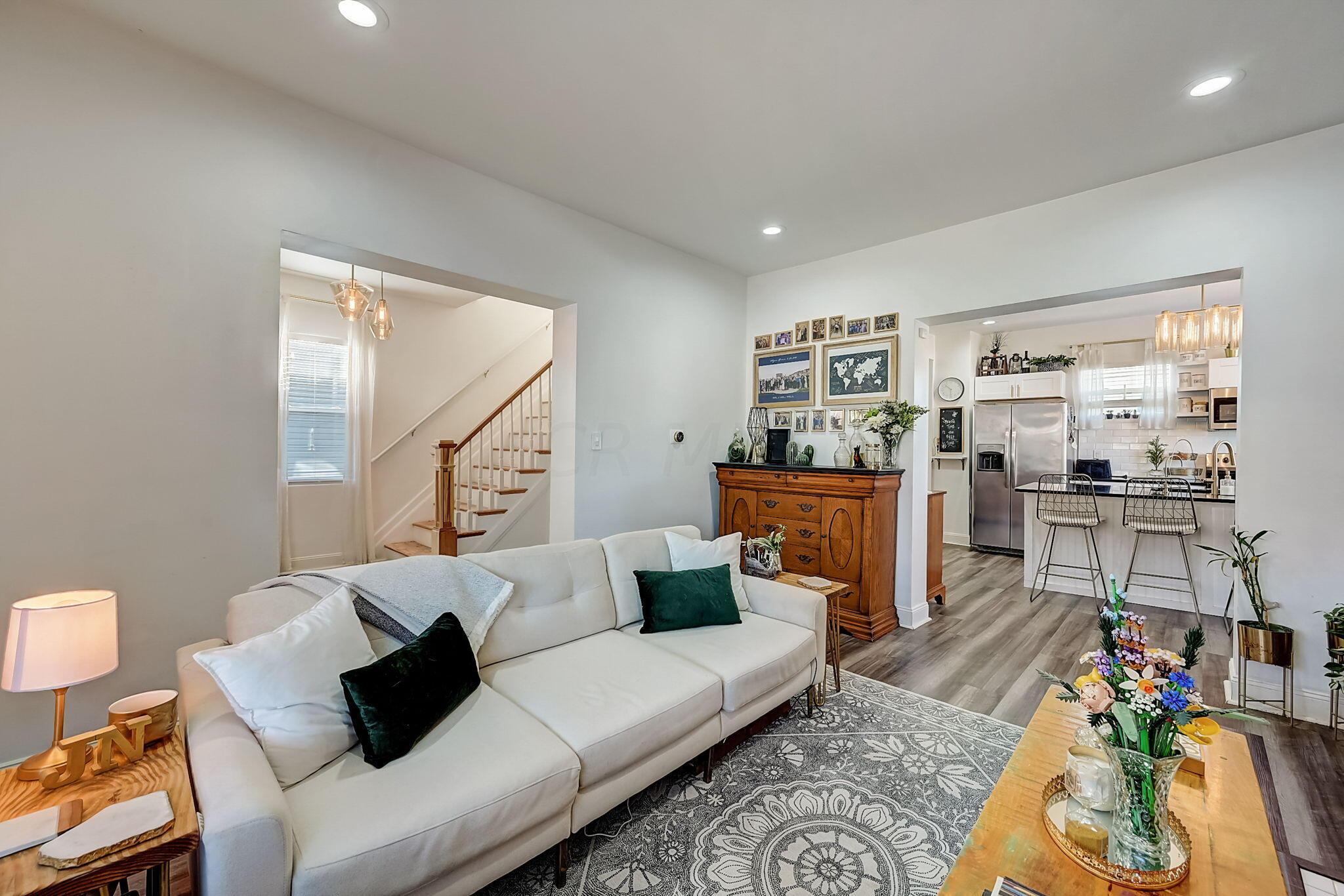 Staged living room with a white sectional, patterned rug, wooden sideboard, and adjoining kitchen.