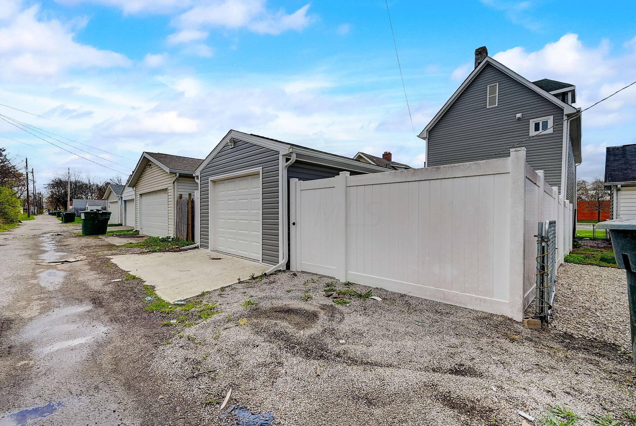 Gray detached garage with a white overhead door beside a tall white privacy fence and gravel alley.
