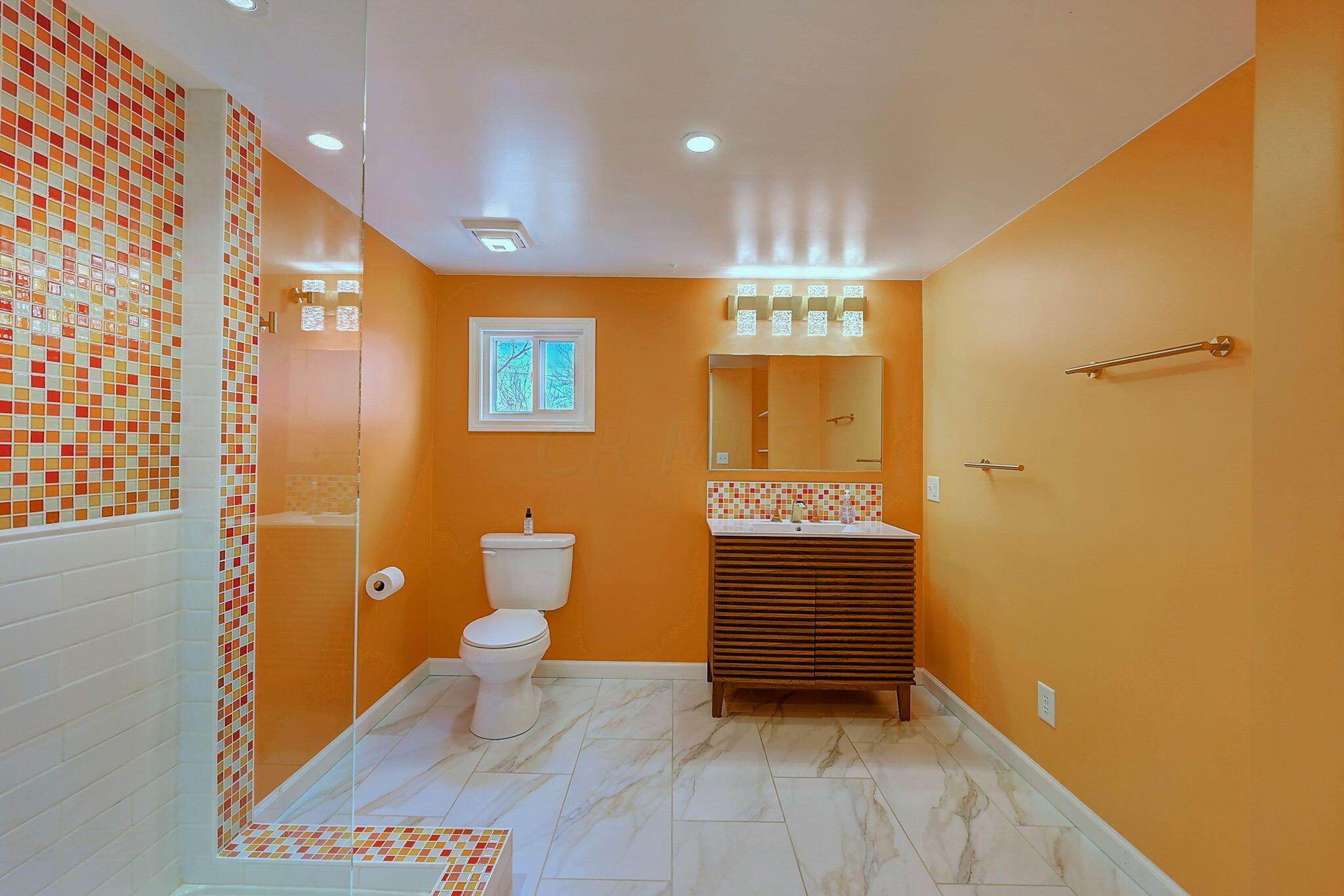 Orange bathroom with a glass-enclosed mosaic-tile shower, white toilet, and wood-slat vanity.