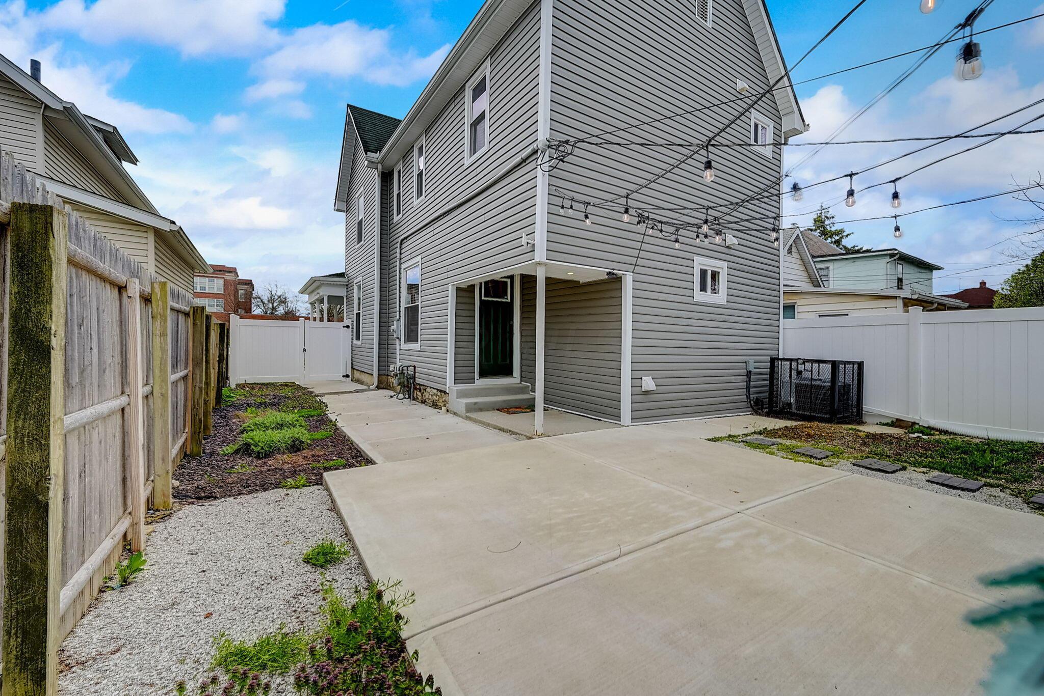 Home’s gray-sided rear exterior with a broad concrete patio, privacy fencing, and string lights.