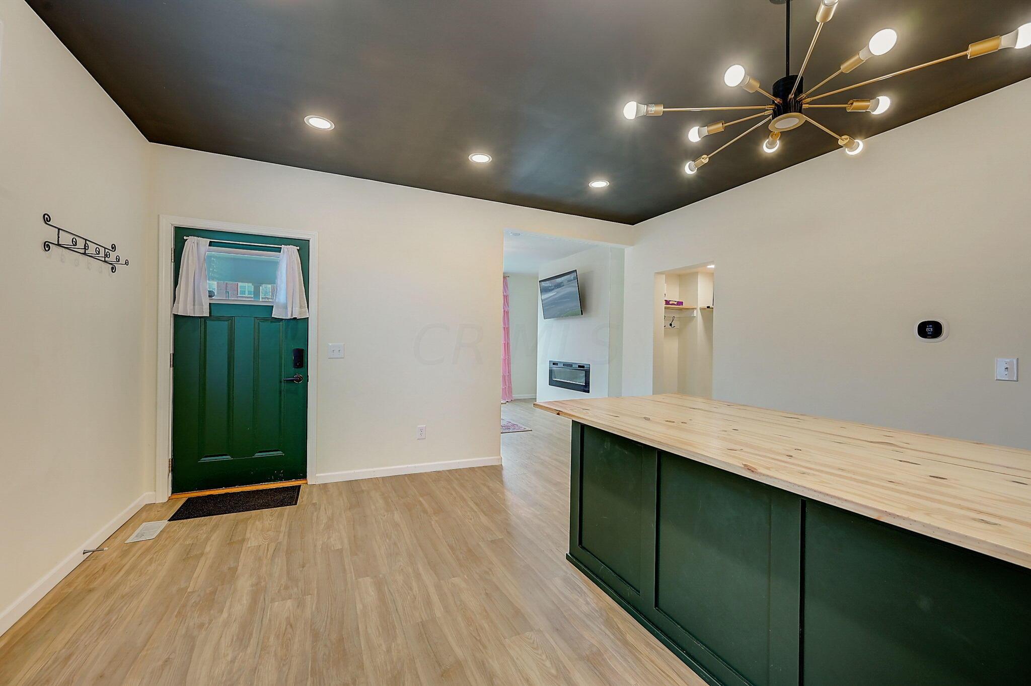Kitchen and entry with a dark green peninsula, light wood countertop, and branching chandelier.