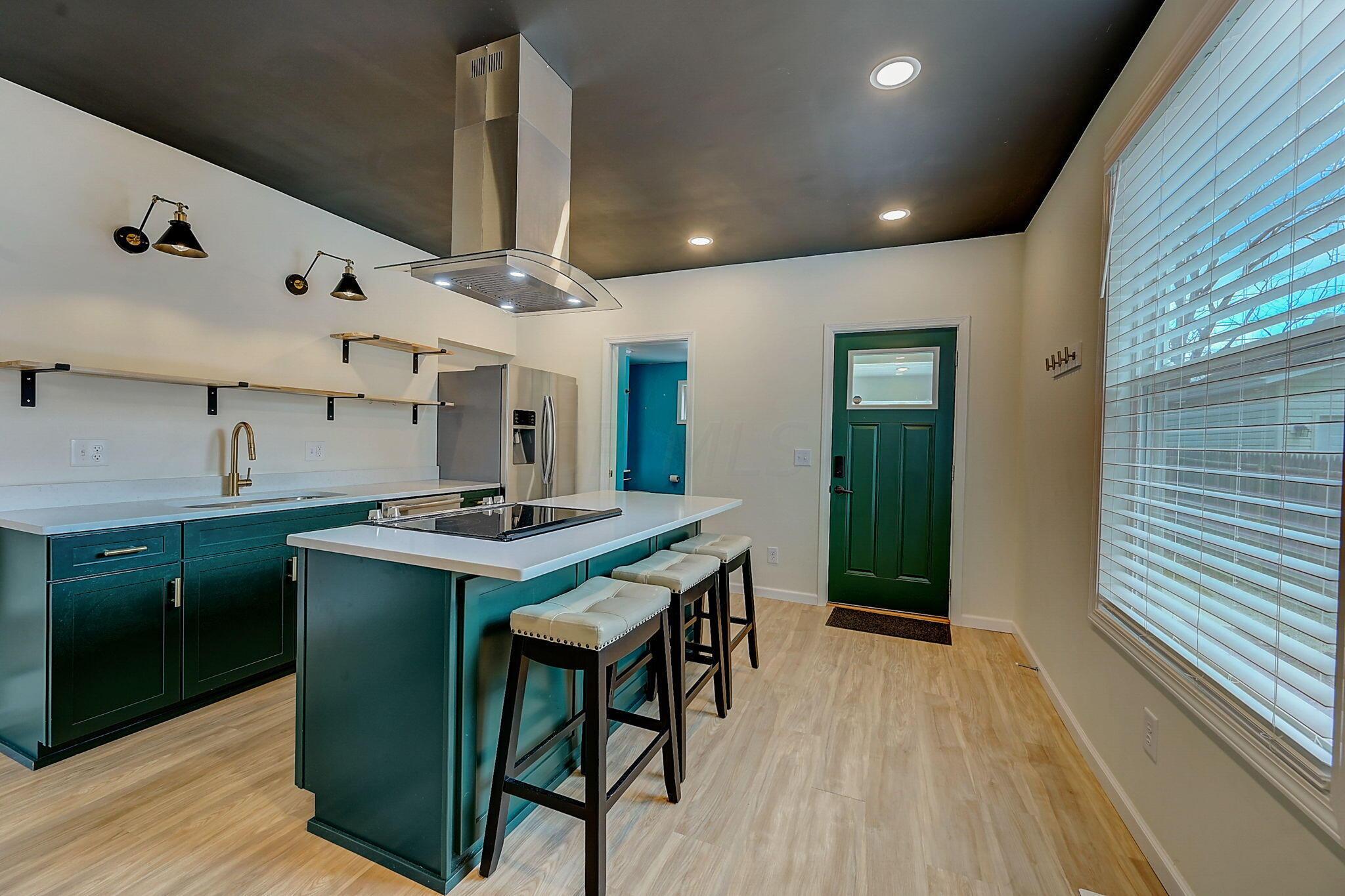 Modern kitchen with teal cabinets, a cooktop island, three bar stools, and a green door.