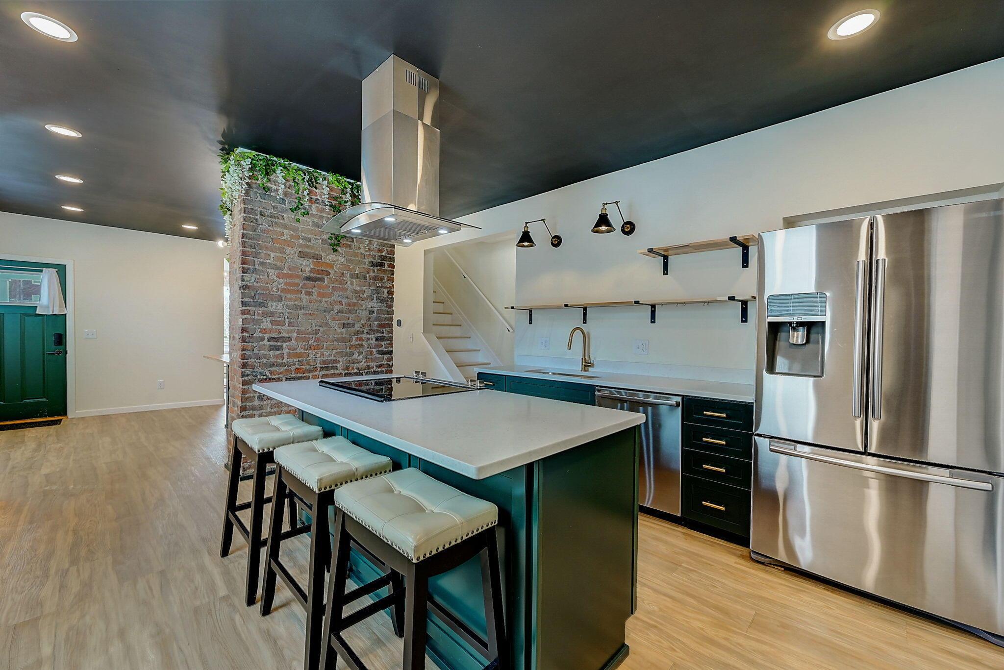 Renovated kitchen with a teal island, three upholstered stools, stainless steel appliances, and an exposed brick wall.