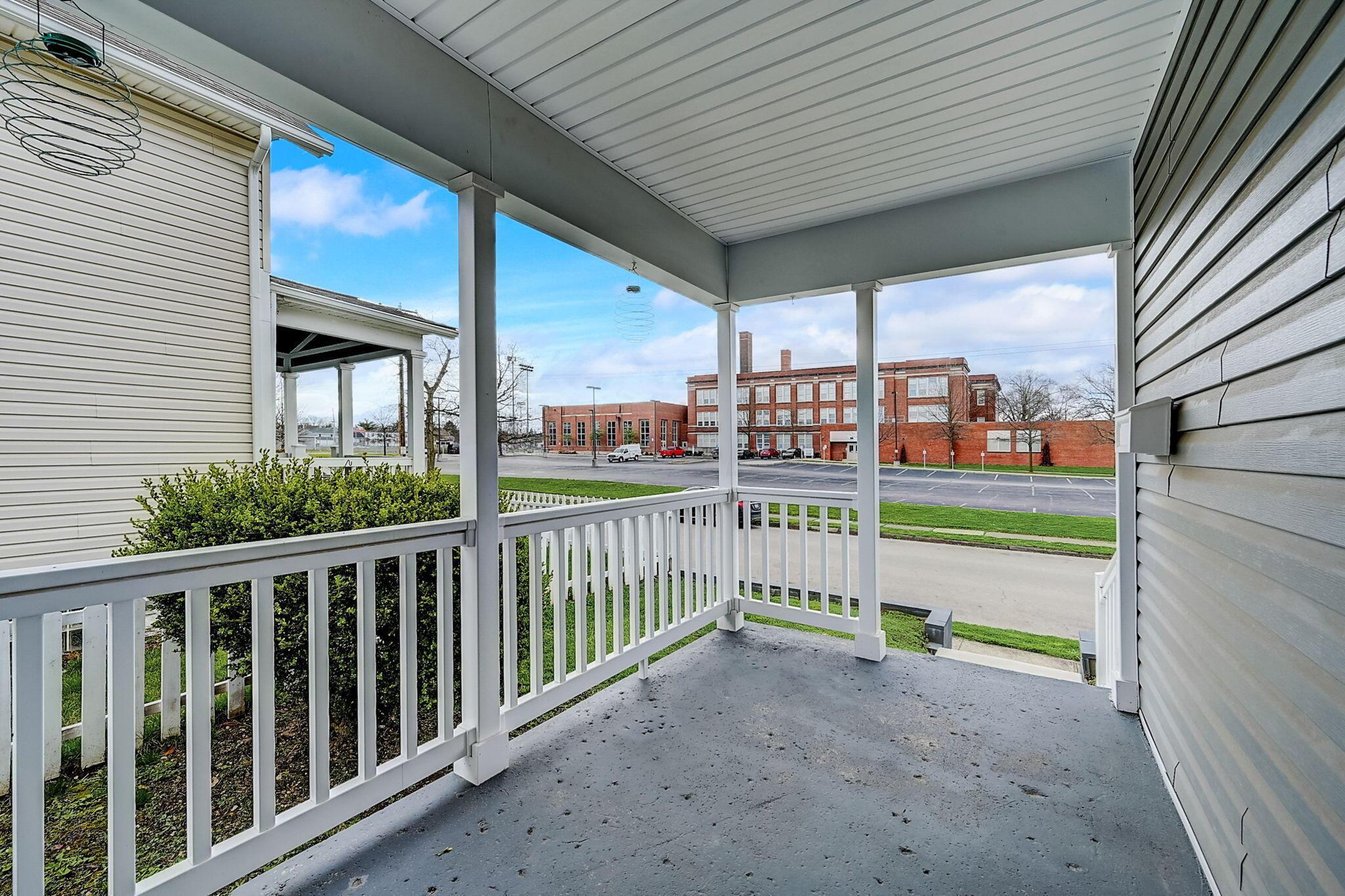 Covered porch with white railings overlooks a street and a large brick building.