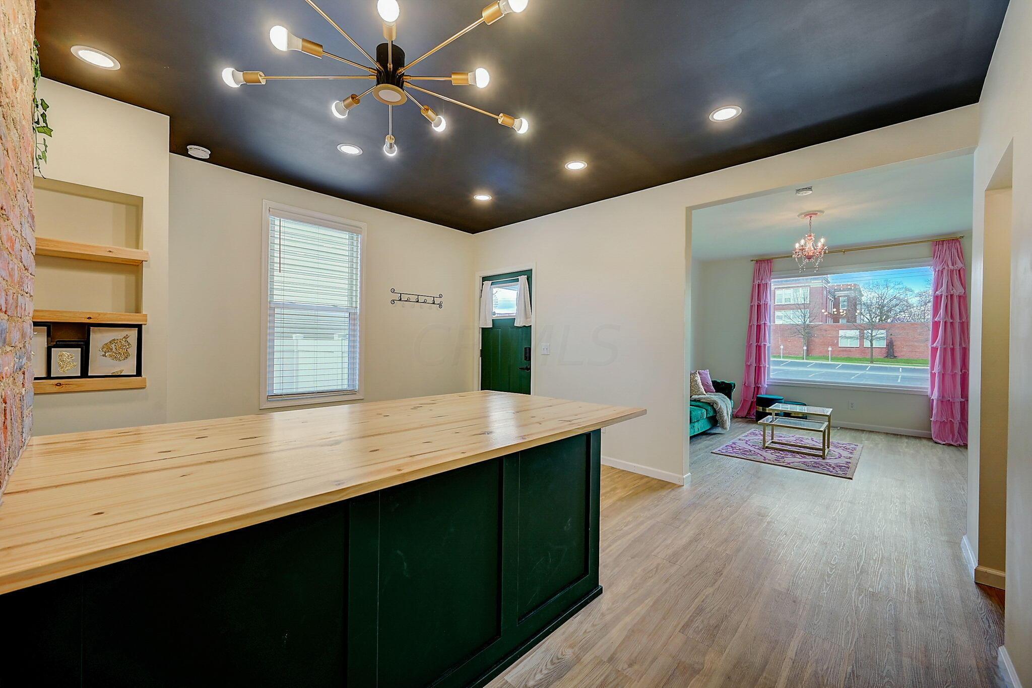 Kitchen with a broad wood-topped green island, opening to a living room with pink curtains.