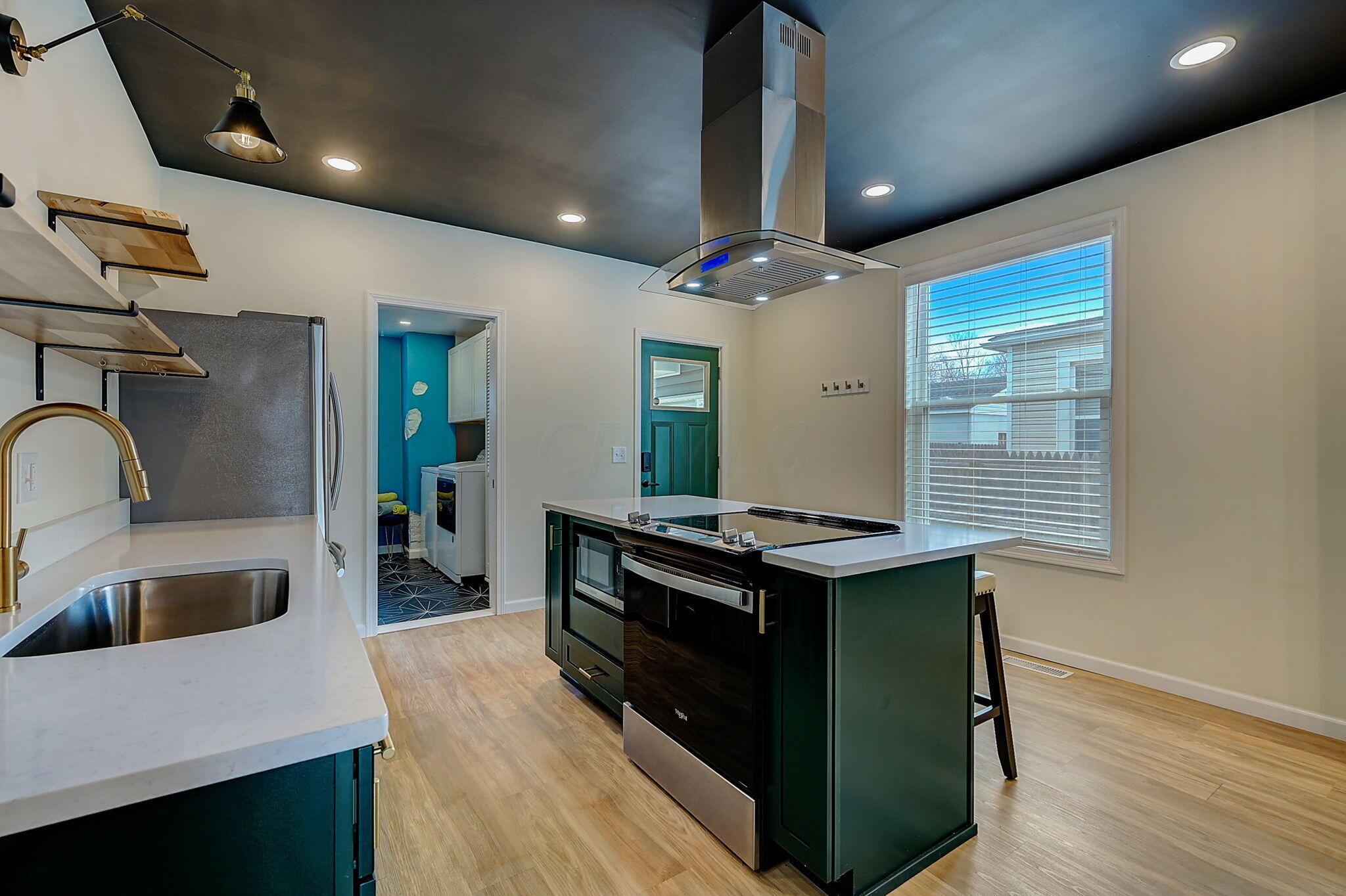 Modern kitchen with a dark green island, white counters, and a stainless range hood.