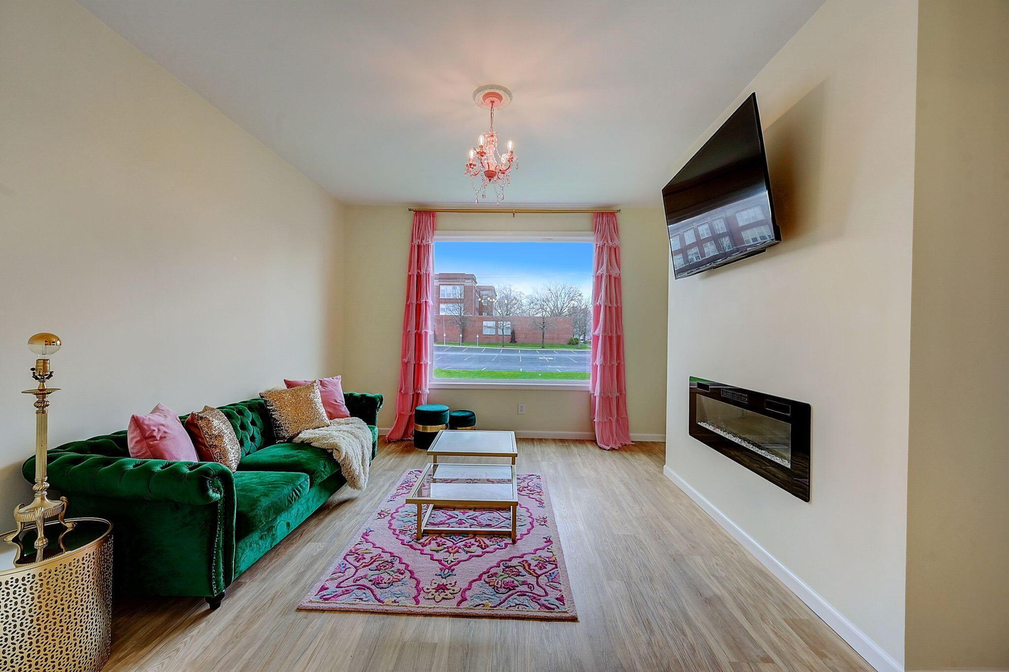 Living room with green tufted sofa, pink curtains, patterned rug, and wall-mounted television above a fireplace.