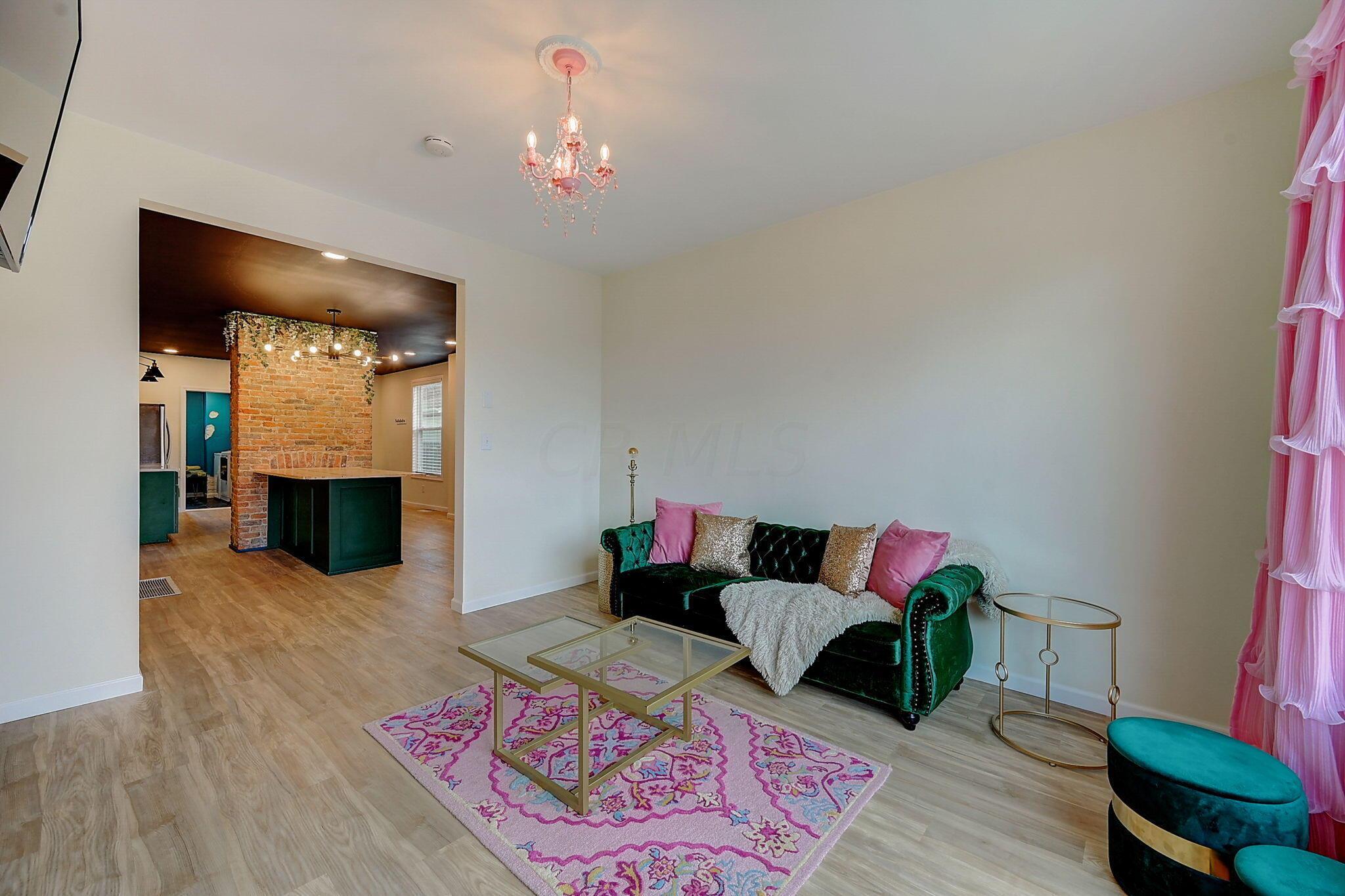 Living room with a green velvet sofa, pink accents, and an adjoining kitchen with an exposed brick column.