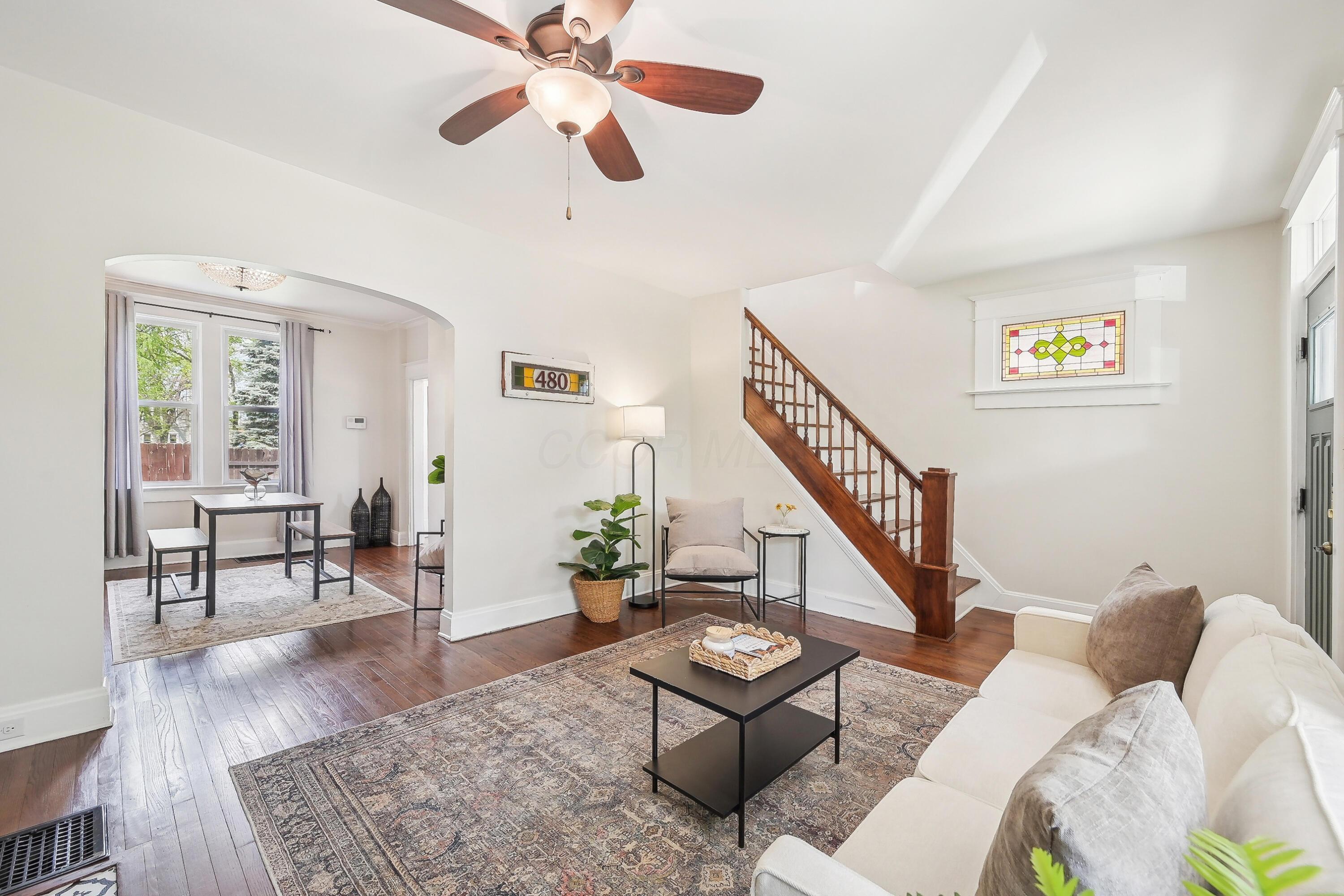 Staged living room with hardwood floors, a wooden staircase, stained-glass window, and arched opening to dining area.