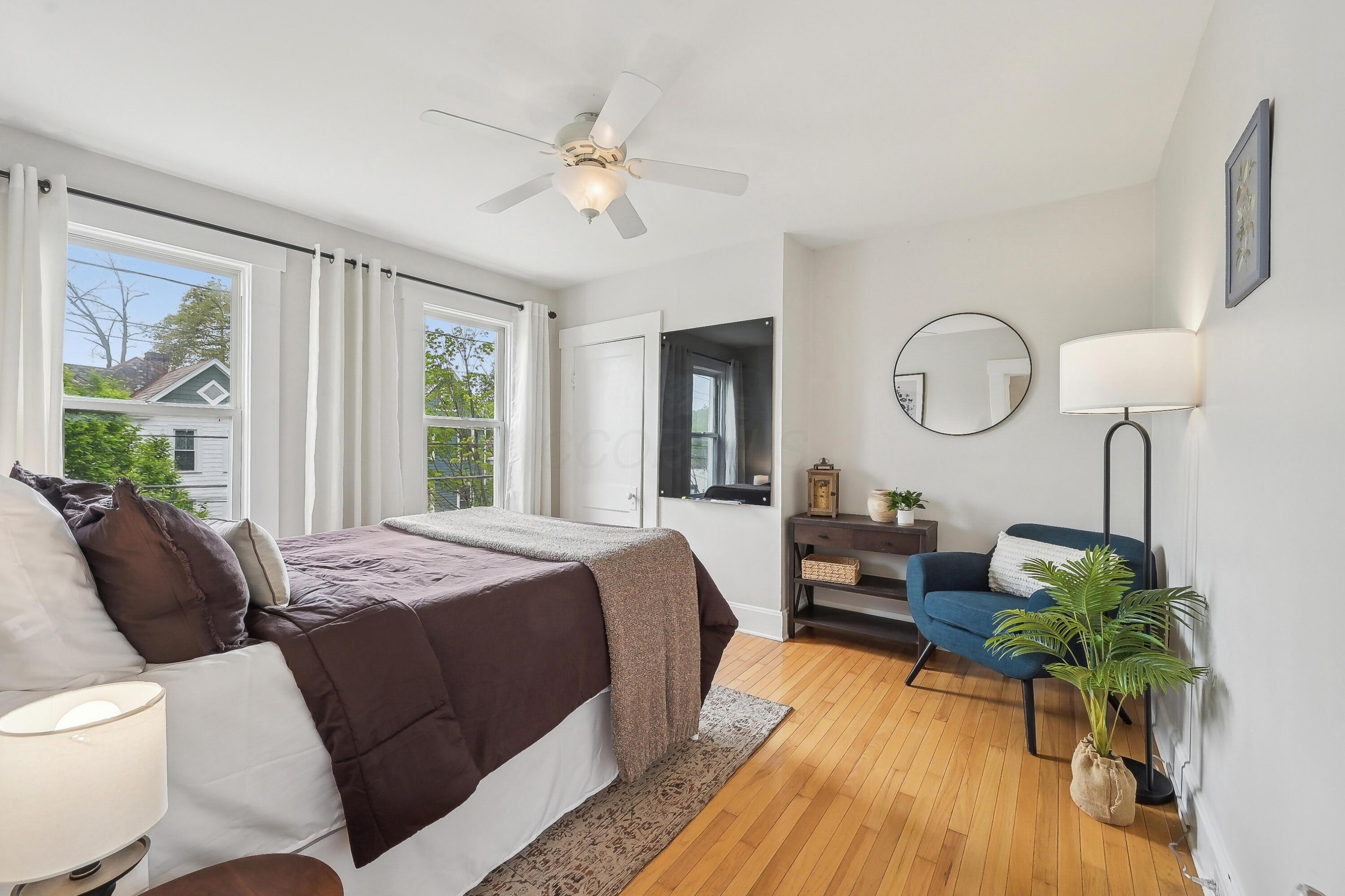 Furnished bedroom with dark plum bedding, hardwood floors, two large windows, and a blue accent chair.