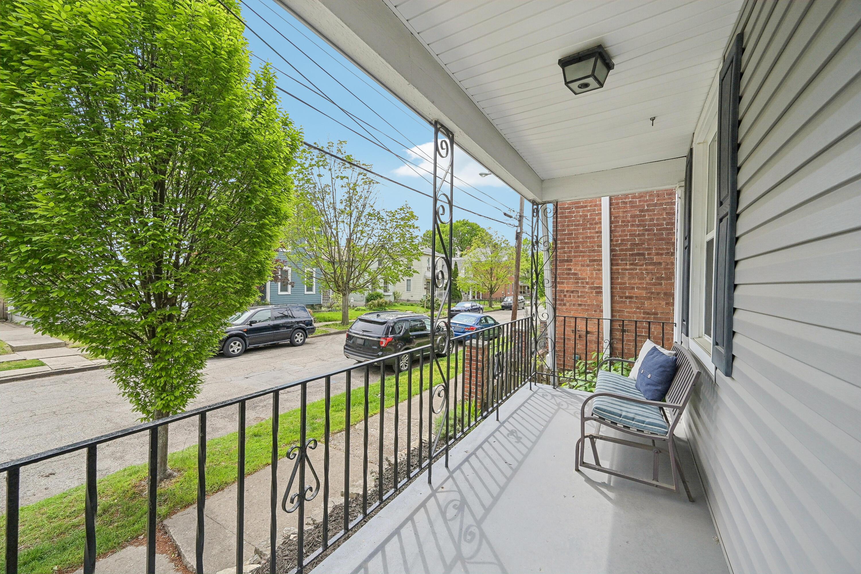 Covered porch with decorative metal railings and a cushioned chair overlooking a tree-lined residential street.