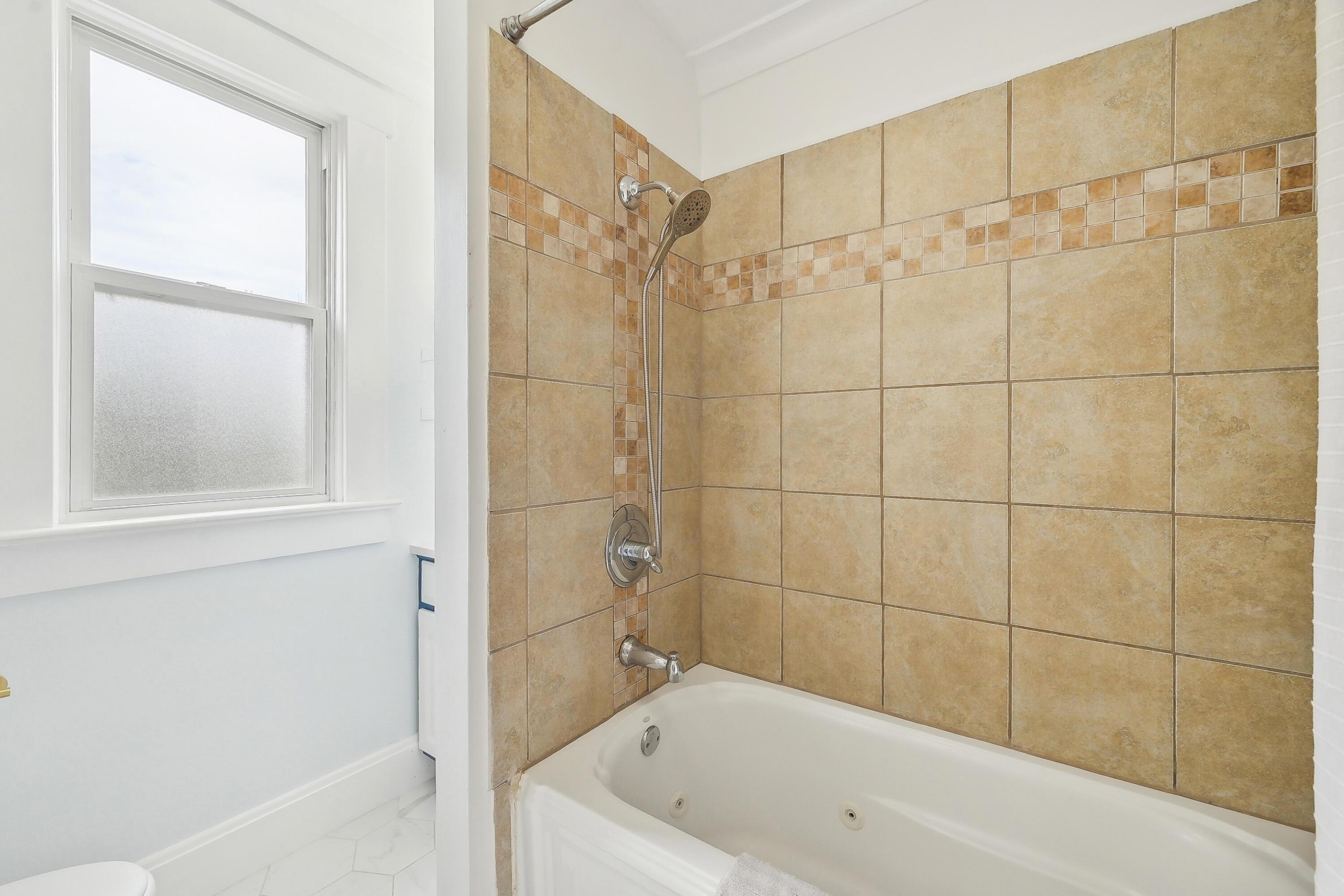 Bathroom bathtub with handheld shower, tan tile surround, and frosted window.