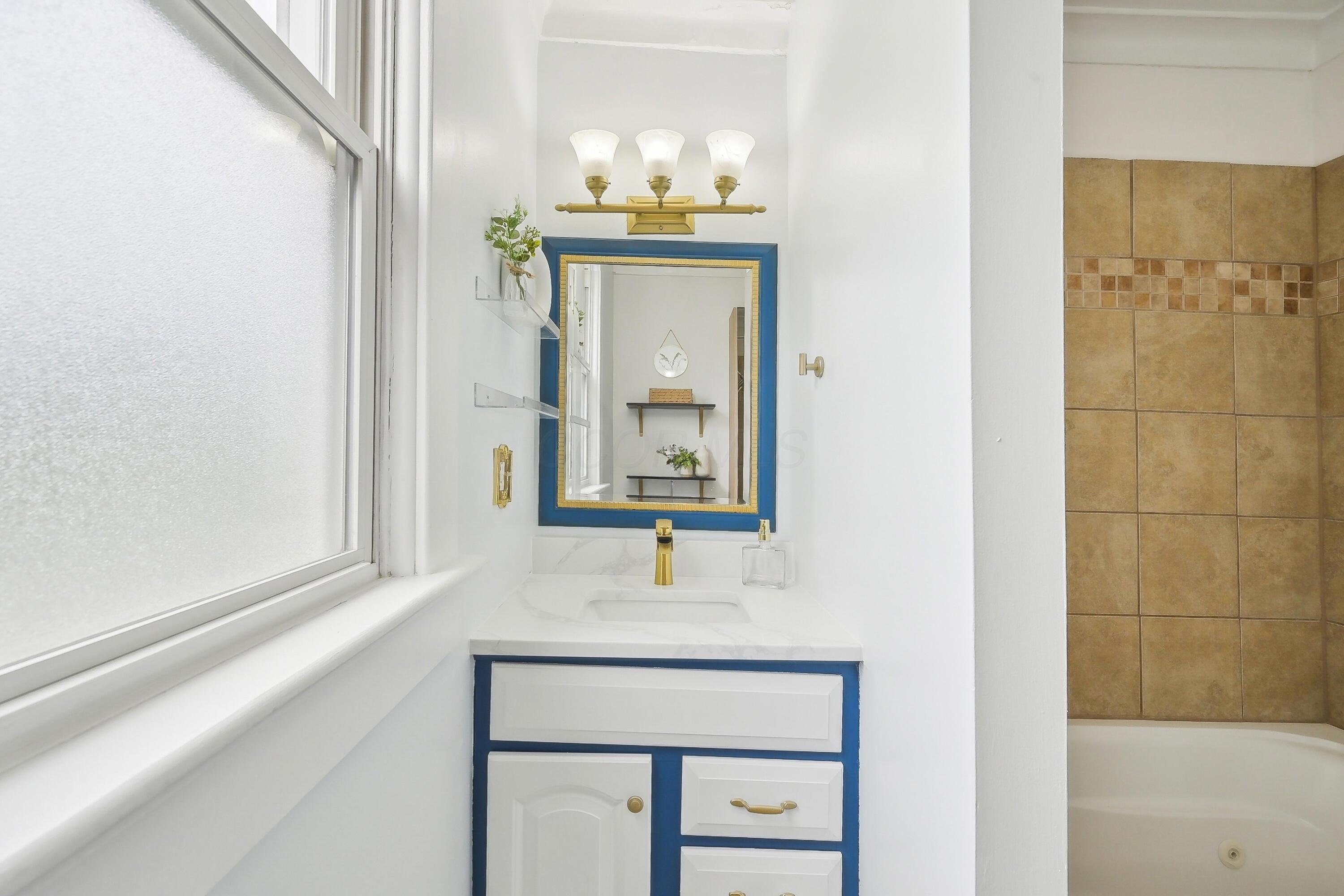 Bathroom vanity with blue-and-white cabinetry, a blue-framed mirror, and gold fixtures beside a large window.