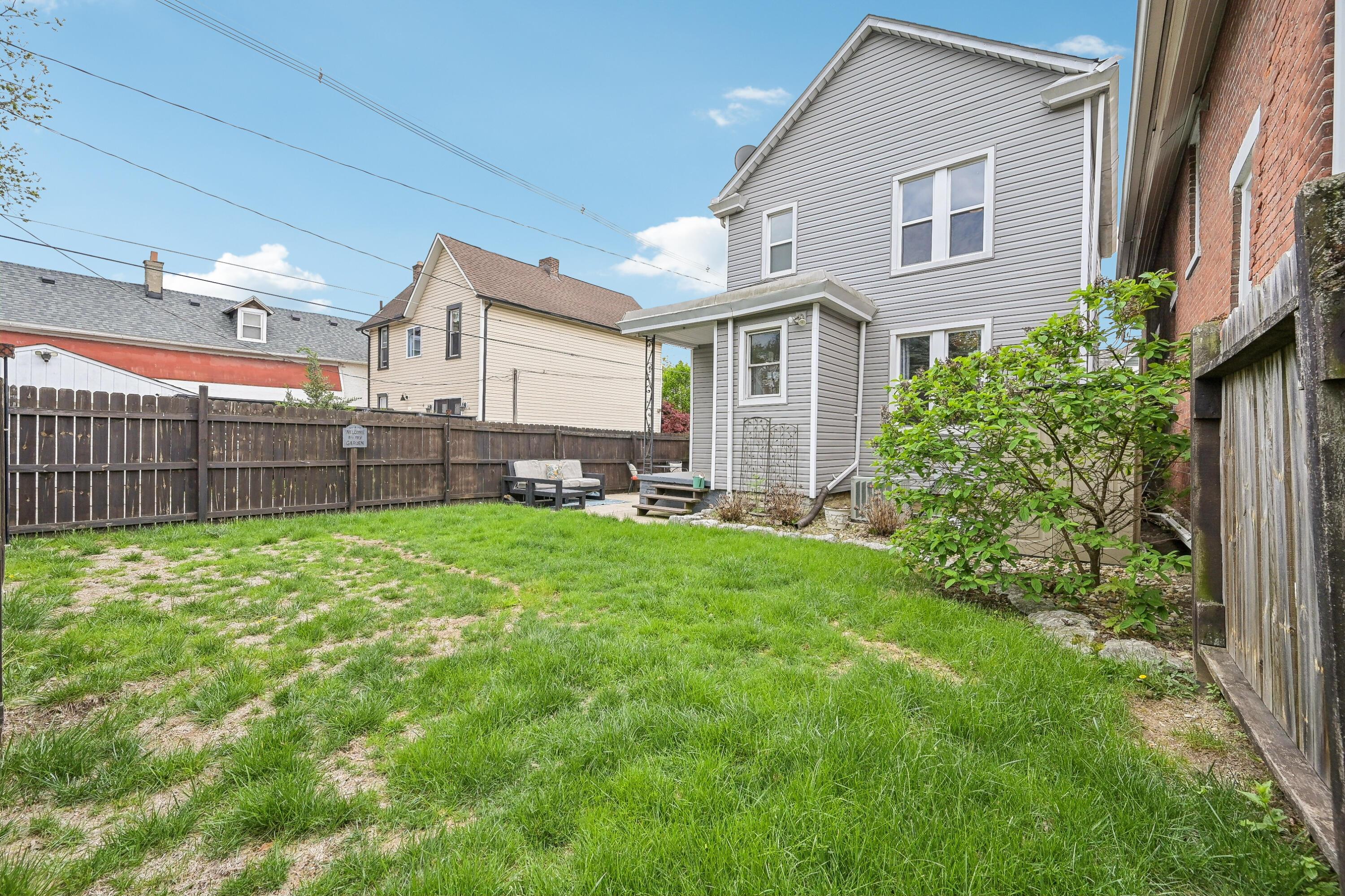 Fenced backyard with lawn, patio seating, and the gray-sided rear of a two-story house.