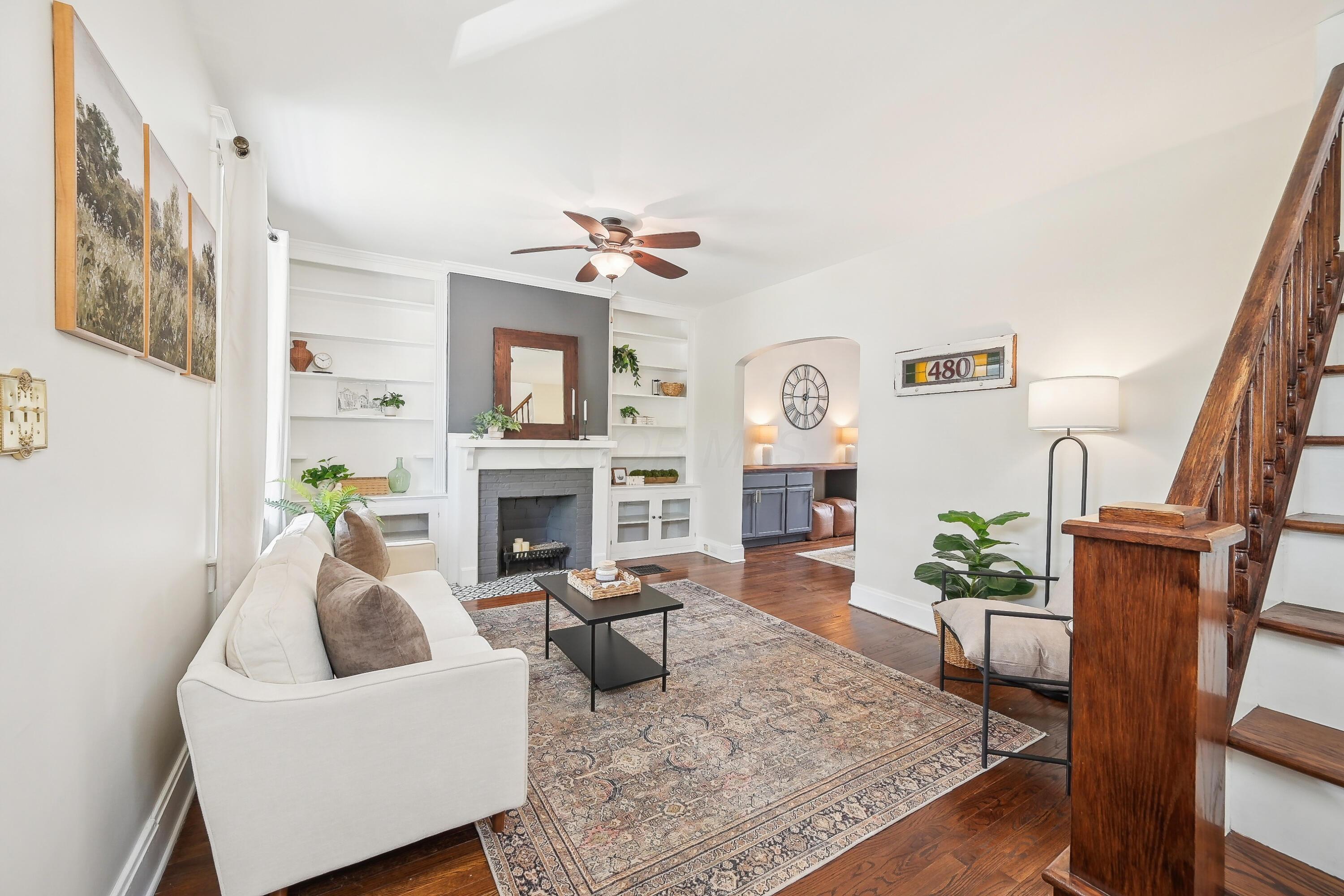 Furnished living room with built-in shelves, a fireplace, patterned rug, and prominent wooden staircase.