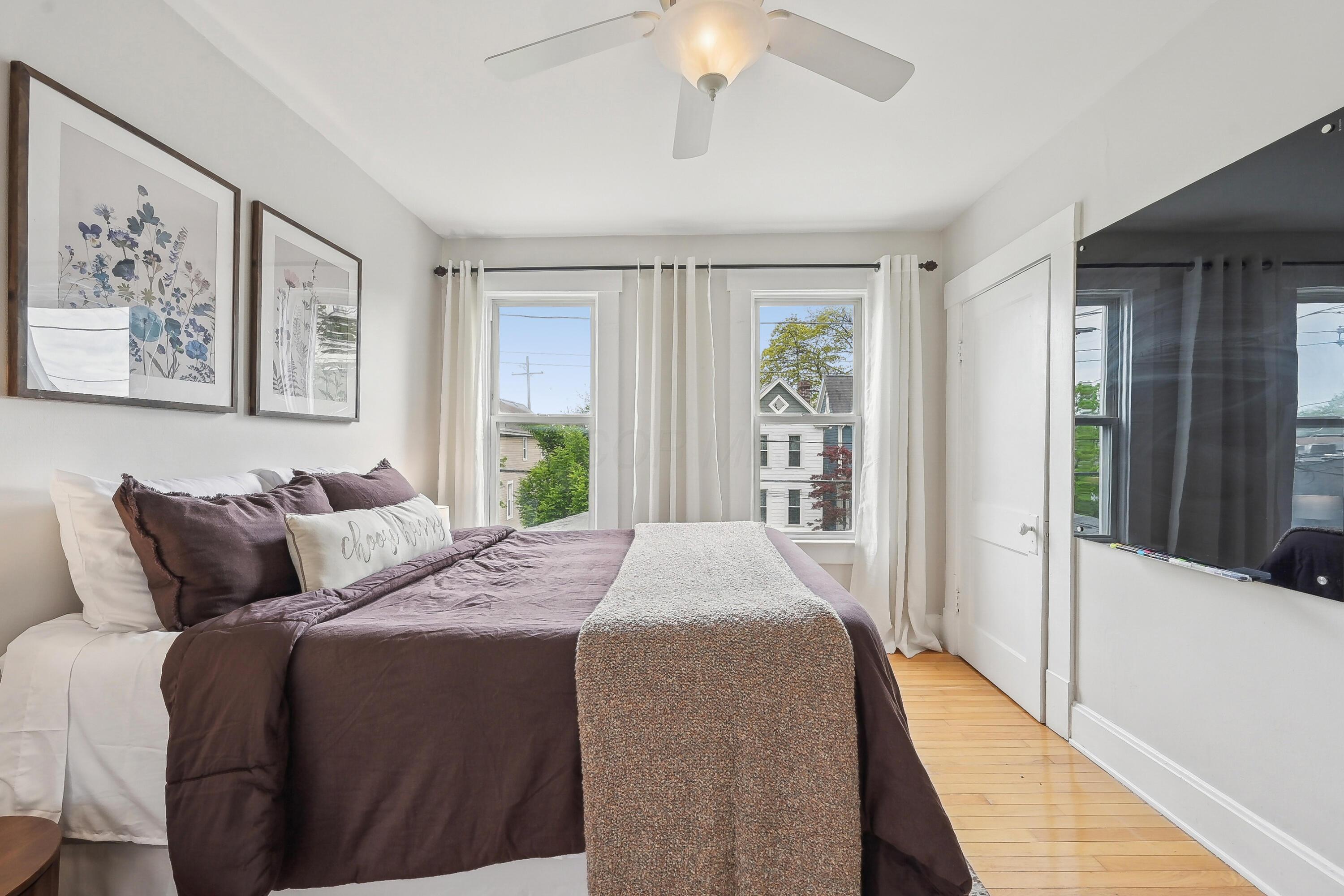 Staged bedroom with plum bedding, botanical prints, and two bright windows.
