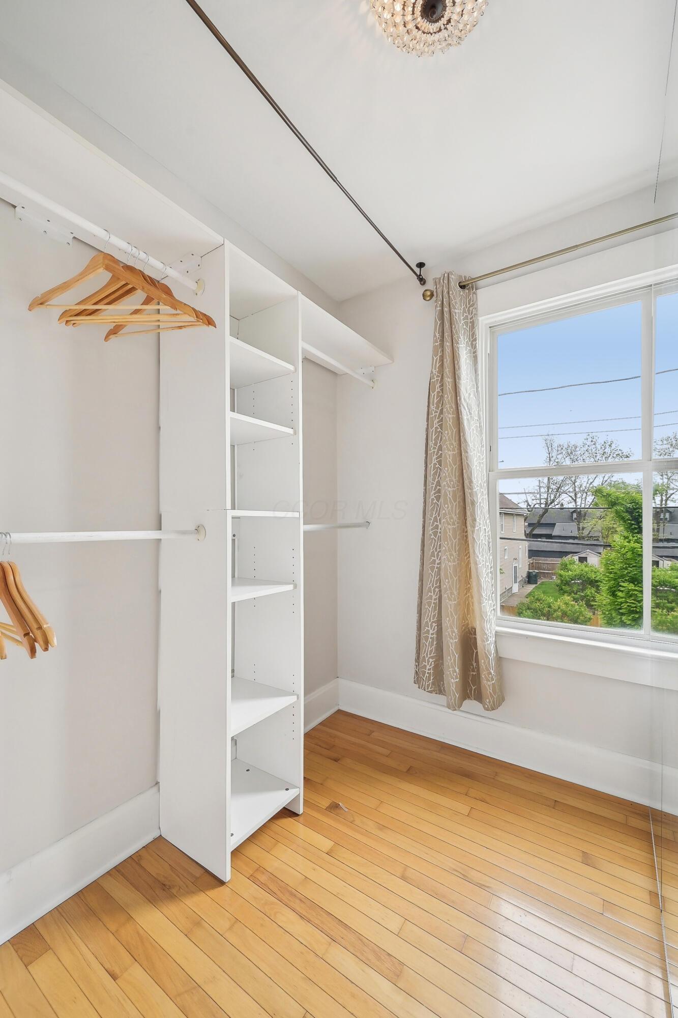 Walk-in closet with white built-in storage, hardwood floors, and a large curtained window.