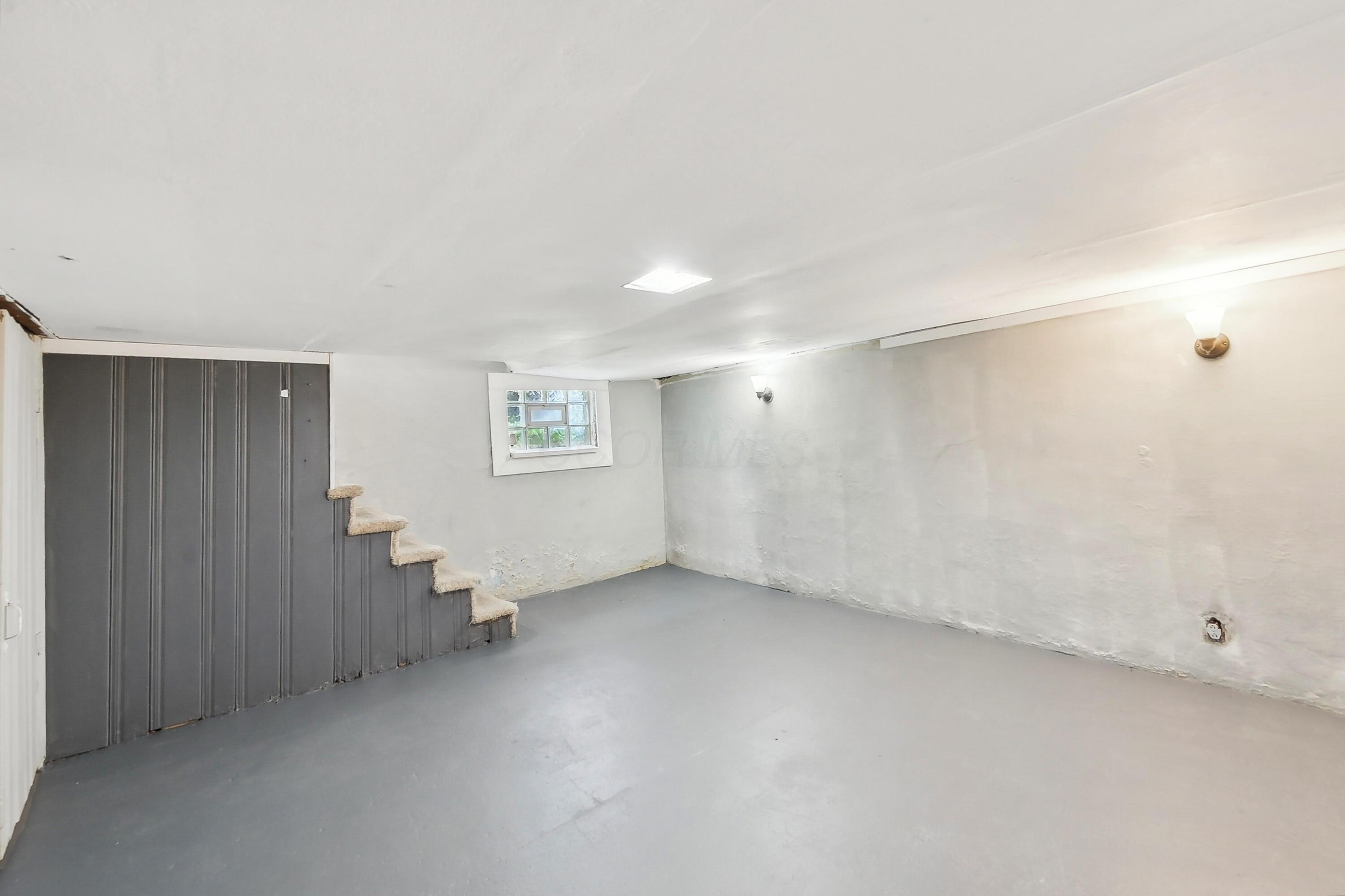 Empty basement with gray flooring, a small window, and carpeted steps beside dark paneled walls.
